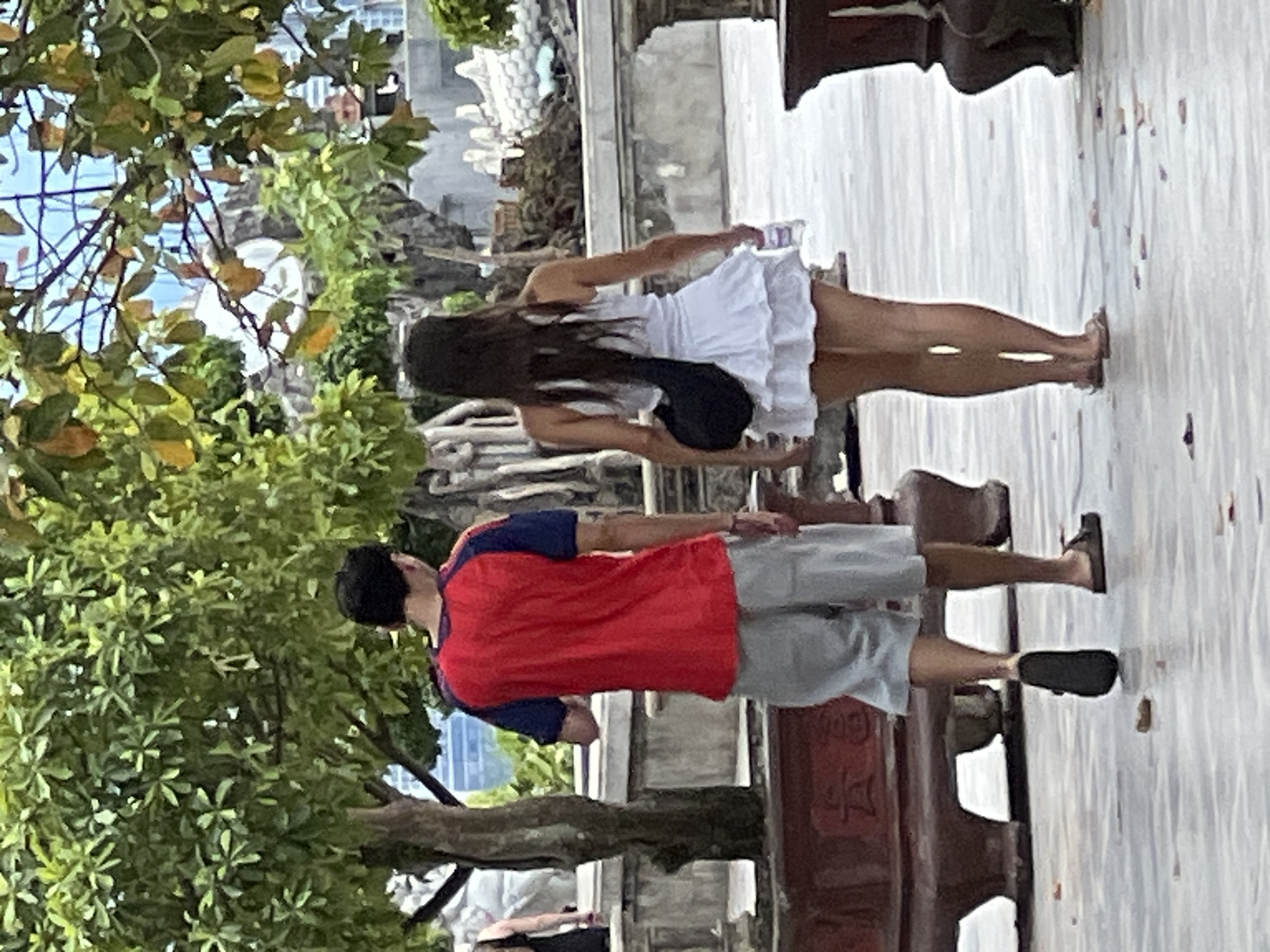 Two individuals, a male and a female, are walking away from the camera on a paved outdoor area in Đà Nẵng, Vietnam. The male is on the left, wearing a red t-shirt with blue sleeves and light gray shorts. He has dark hair and is wearing dark footwear. The female is on the right, wearing a white sleeveless ruffled dress and carrying a black bag over her shoulder. She has long dark hair and is wearing light-colored sandals. Both are walking on a light-colored paved surface, possibly stone or concrete, which shows some wetness and scattered debris, likely leaves or flower petals. In the background, there are trees with lush green foliage, some with dried leaves. Behind the trees, architectural elements suggestive of a traditional Asian structure, possibly a temple or pagoda, are visible, including ornate carvings and roofing details. The overall environment appears to be a public space or garden area within the city.