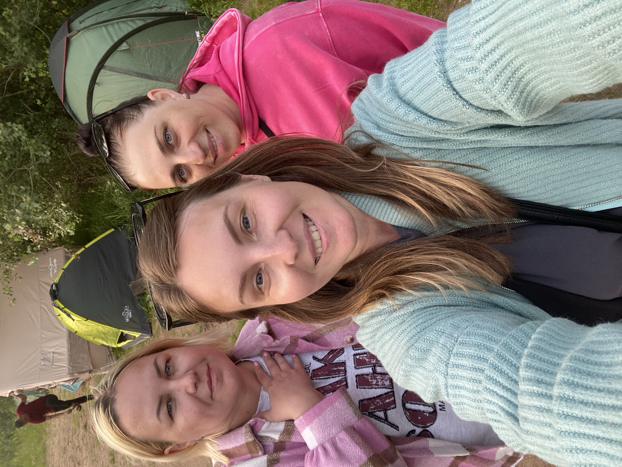 The image shows three women in a selfie taken outdoors, likely during a camping trip. Two tents are visible in the background. One is a green dome tent, and the other is a larger tan tent. The women are smiling and appear to be in good spirits.
The woman in the foreground has blonde hair and is wearing a pink and white plaid jacket over a t-shirt with a graphic print. Her left hand is resting on her chest. Next to her, slightly above, is a woman with long brown hair, wearing a light blue knitted sweater. She is also smiling. The third woman, in the background, has dark hair pulled back and is wearing a bright pink hooded sweatshirt. She is wearing sunglasses on her head.
The lighting suggests it is daytime, possibly late afternoon or early evening, given the soft light. The setting appears to be a grassy area with trees and foliage in the background, consistent with a campsite. There is no visible text in the image. The overall mood is cheerful and relaxed, capturing a moment of friendship and outdoor recreation. The location context is provided as Ольховка, Russia.