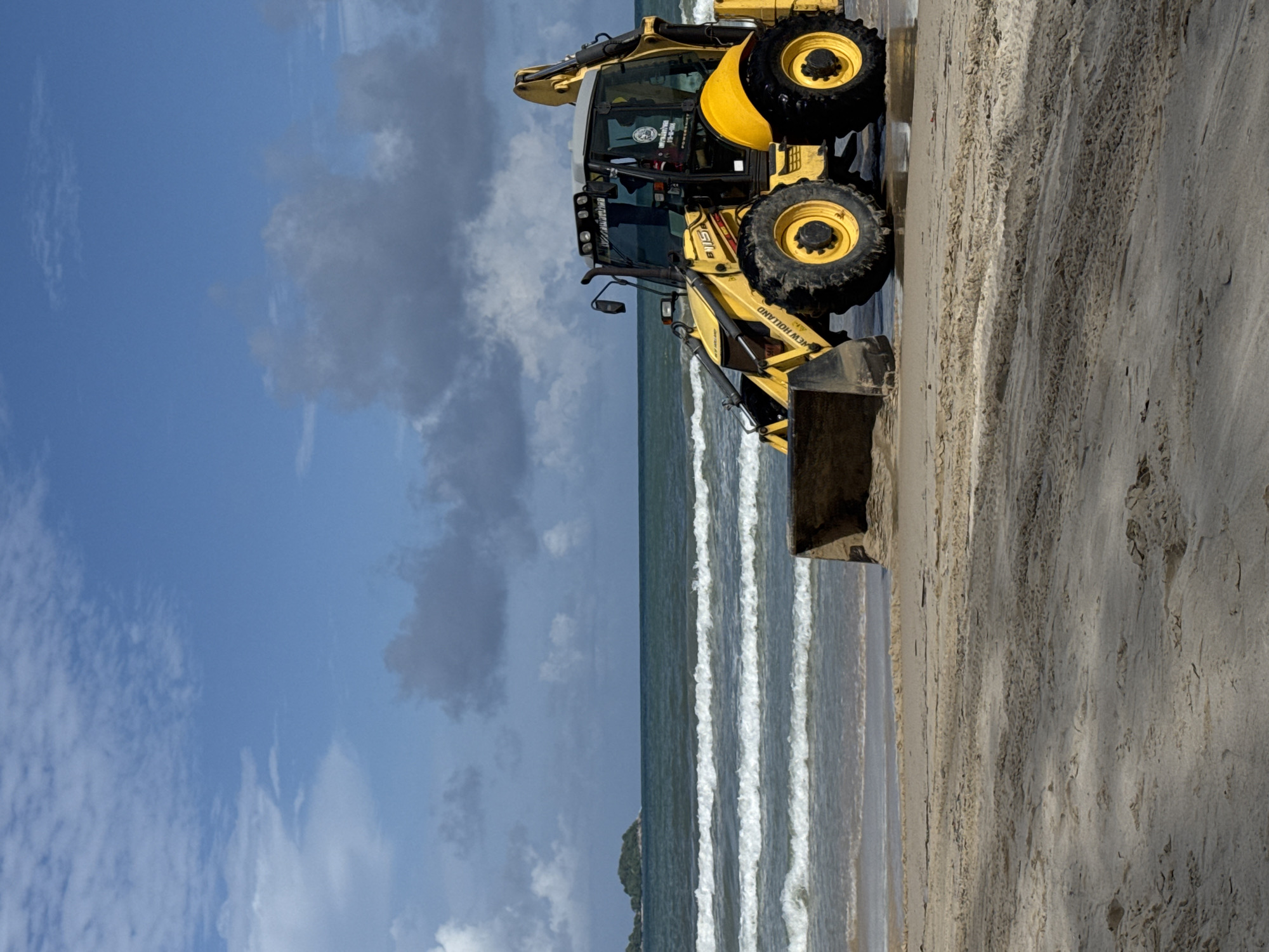 A yellow New Holland backhoe is parked on the wet sand of Patong Beach, Thailand. The machine is facing right, with its front loader lowered to the ground. Waves are gently lapping at the shore behind the vehicle, and a small, tree-covered island is visible in the distance to the left. The sky above is blue with scattered white and grey clouds. The sand is damp and marked with tire tracks.