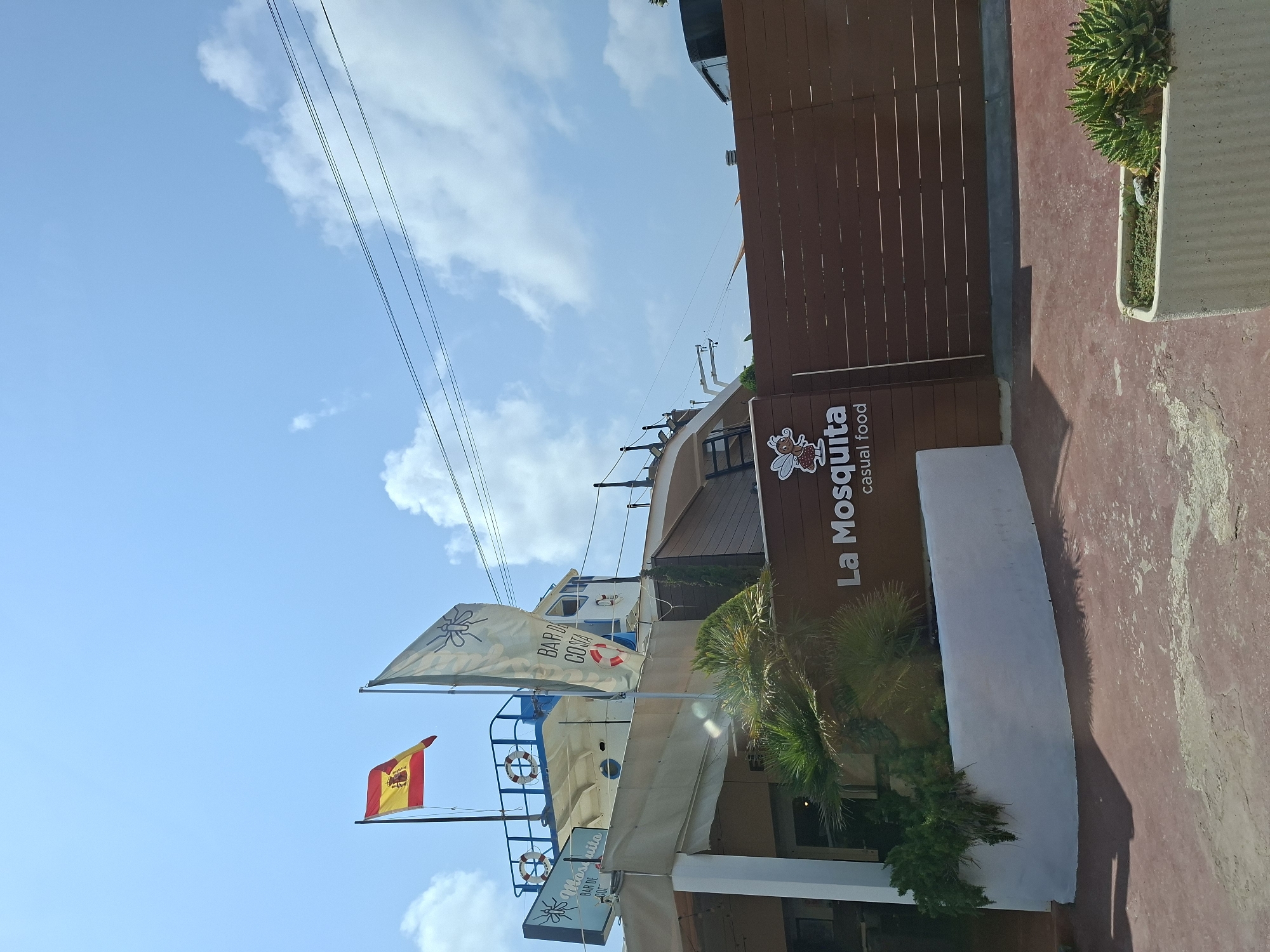 The image shows a building with a distinctive boat-like structure on top, featuring the Spanish flag and a white flag with a mosquito logo. Signs indicate "Mosquito Bar de Costa" and "La Mosquita casual food". The building has a light-colored facade with a large awning. In the foreground, a dark wooden fence runs horizontally across the frame, and a concrete planter with succulents is visible to the right. The ground is paved with a reddish material. The sky is blue with scattered white clouds, and power lines are present in the upper right corner. This scene is located in Cabo De Palos, Spain.
