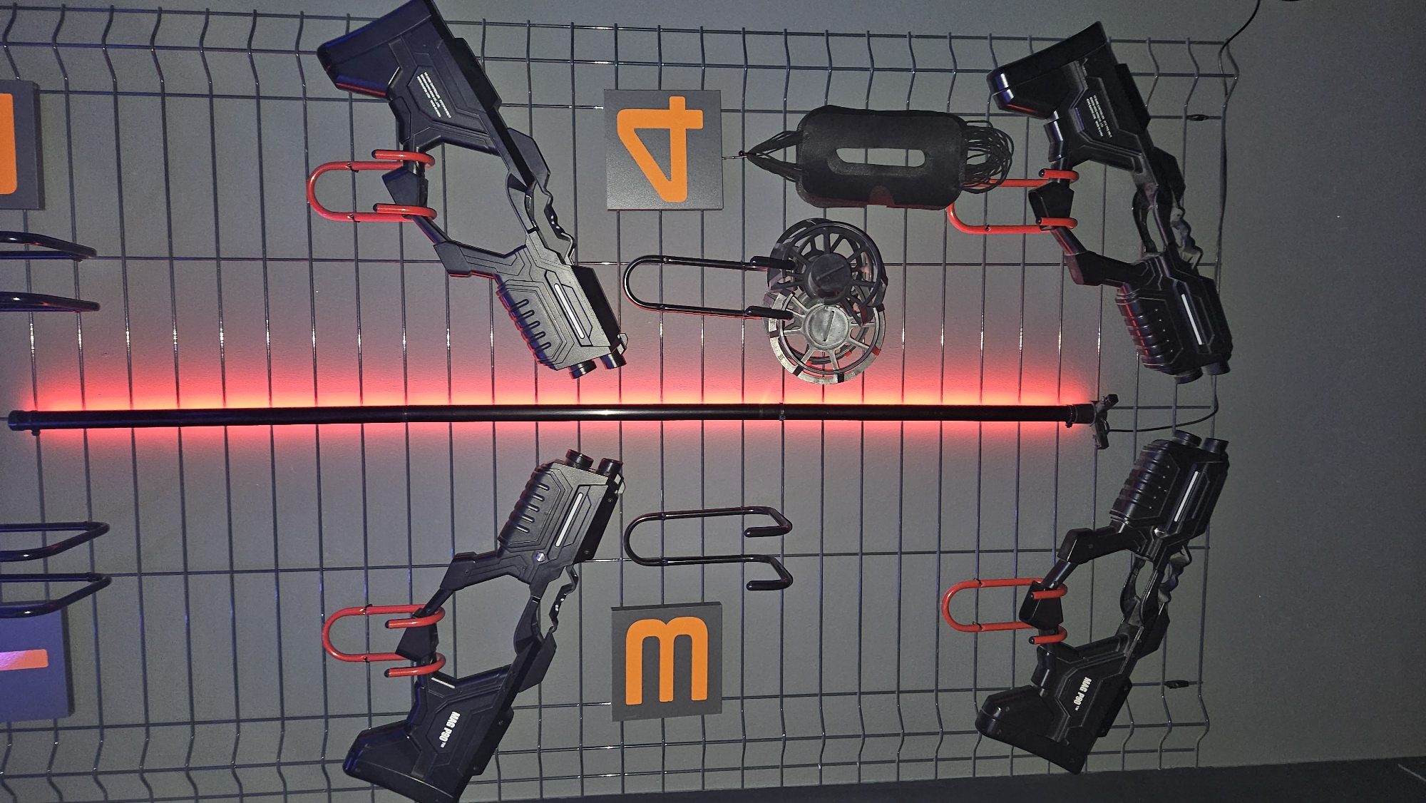 A wall display in an indoor setting features several items mounted on a wire grid. A vertical, thin, black pole with a red light emanating from it runs down the center of the image. On the left side of the display, two black, futuristic-looking handguns are mounted horizontally by red hooks. Below these, a square orange sign with the number "3" is visible, and beneath that, a black double-hook rack. To the right of the central pole, two more identical handguns are mounted, also by red hooks. Below the right-hand handguns, a square orange sign with the number "4" is displayed. To the right of the "4" sign, a black, soft, rectangular object hangs vertically, possibly a punching bag or weighted item. To the left of the "4" sign and below the handguns, a circular metallic object with fan-like blades is suspended. The overall impression is a display for a game or activity involving these prop weapons and accessories.