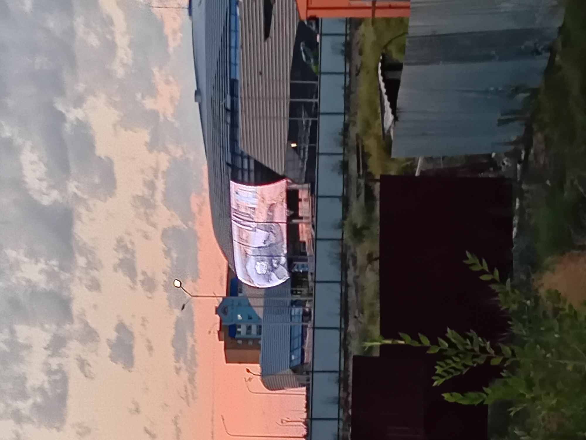 The image shows a partially constructed or renovated building with a curved, illuminated screen displaying an image. The building has a modern architectural design with curved roofs and a facade of glass and metal panels. In the background, several other buildings are visible, including a taller residential building with blue and orange accents. The sky is a mix of pale pink and gray clouds, suggesting either sunrise or sunset. The foreground shows a dark, rectangular structure, possibly a fence or wall, with some greenery in front of it. A corrugated metal fence runs across the middle ground. The scene appears to be in an urban or industrial area, possibly a construction site. The location is identified as Semey, Kazakhstan.