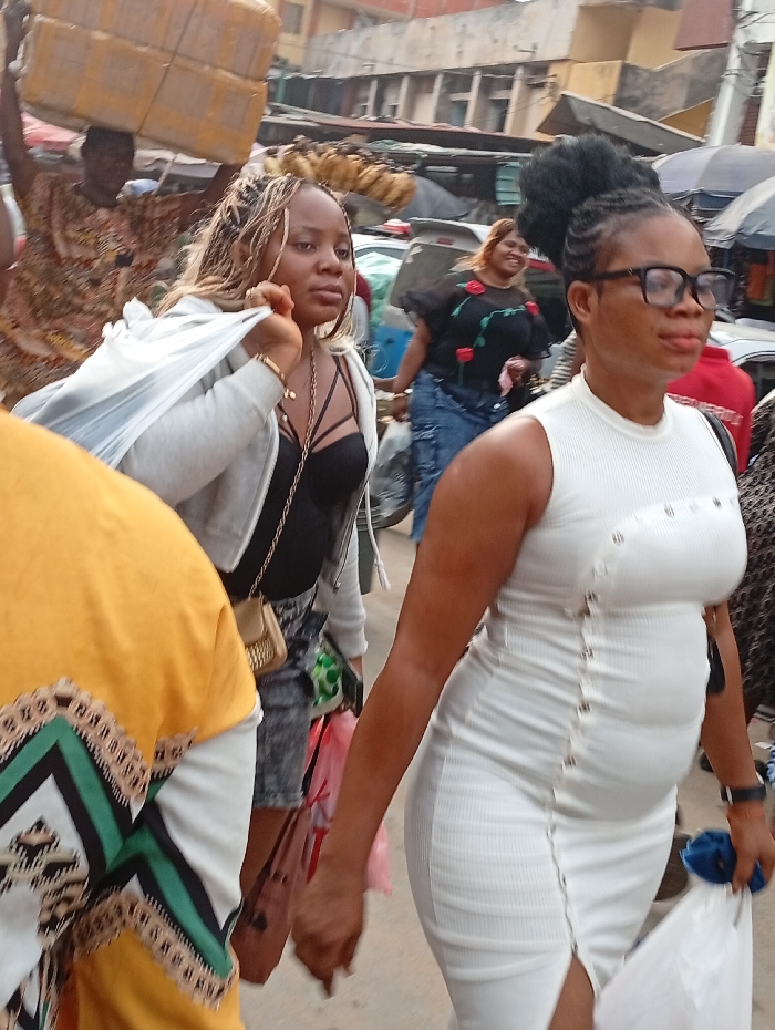 Two women walk side by side in a crowded market. The woman on the left has blonde highlights, a black top, a gray jacket, and denim shorts. The woman on the right wears a white dress and glasses. A man in the background carries a large stack of boxes on his head. Another woman in a black floral top and jeans stands behind them, smiling. The market is busy with people, cars, and goods for sale.