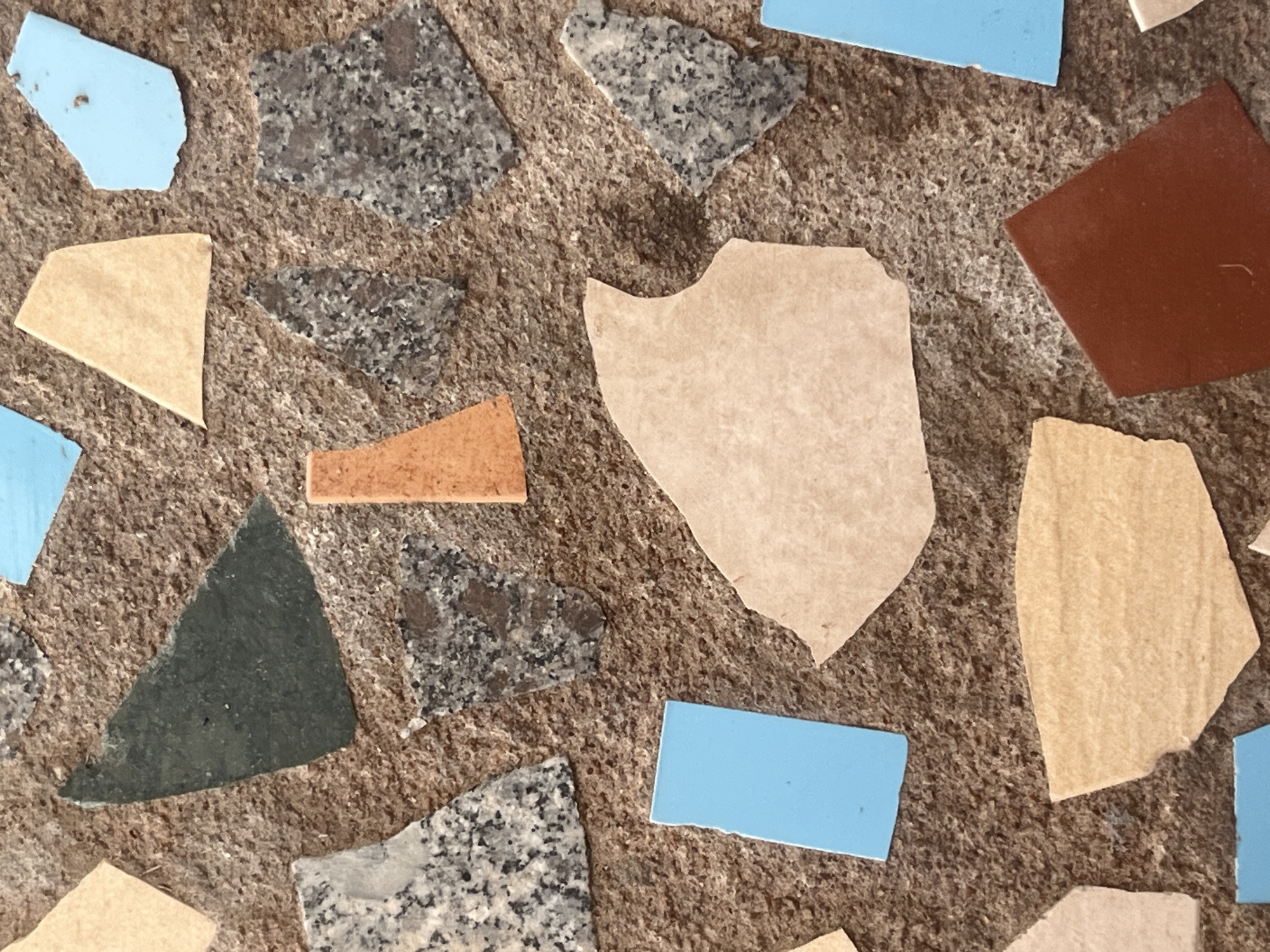 This is a close-up, overhead shot of a mosaic-style surface. The surface is textured and appears to be made of small, irregularly shaped pieces of various materials embedded in a darker, rougher base. The embedded pieces include fragments of what look like granite or speckled stone in grey and black, some light tan or beige pieces, bright blue flat rectangular pieces, a dark green triangular piece, and a small, angular orange piece. The overall composition suggests an artistic or decorative element, possibly a pathway, wall, or tabletop. There is no indication of a specific location within Mũi Né, Vietnam; this could be any decorative surface. There are no people or discernible actions taking place. The lighting is even, suggesting it was taken during the day, likely outdoors or in a well-lit indoor space.