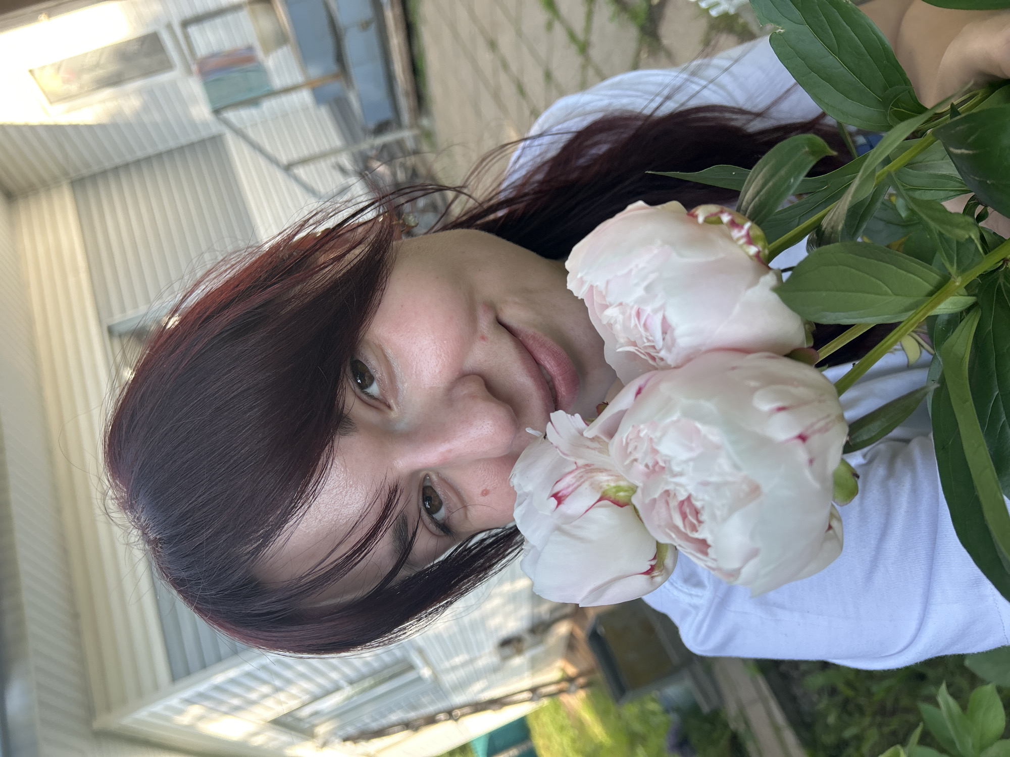 A medium shot captures a young woman with dark hair and fair skin holding a bouquet of light pink peonies. She is positioned outdoors, likely in a yard or garden, as suggested by the presence of greenery and what appears to be a house in the background with horizontal siding. The woman is wearing a white t-shirt and looking directly at the camera with a gentle smile. Her face is illuminated by natural light, highlighting her features. The peonies are the focal point alongside her face, with their delicate petals and hints of red at the edges. The background is somewhat blurred, but hints at a residential setting, possibly in the city of Fokino, Russia, given the architectural style that can be observed.