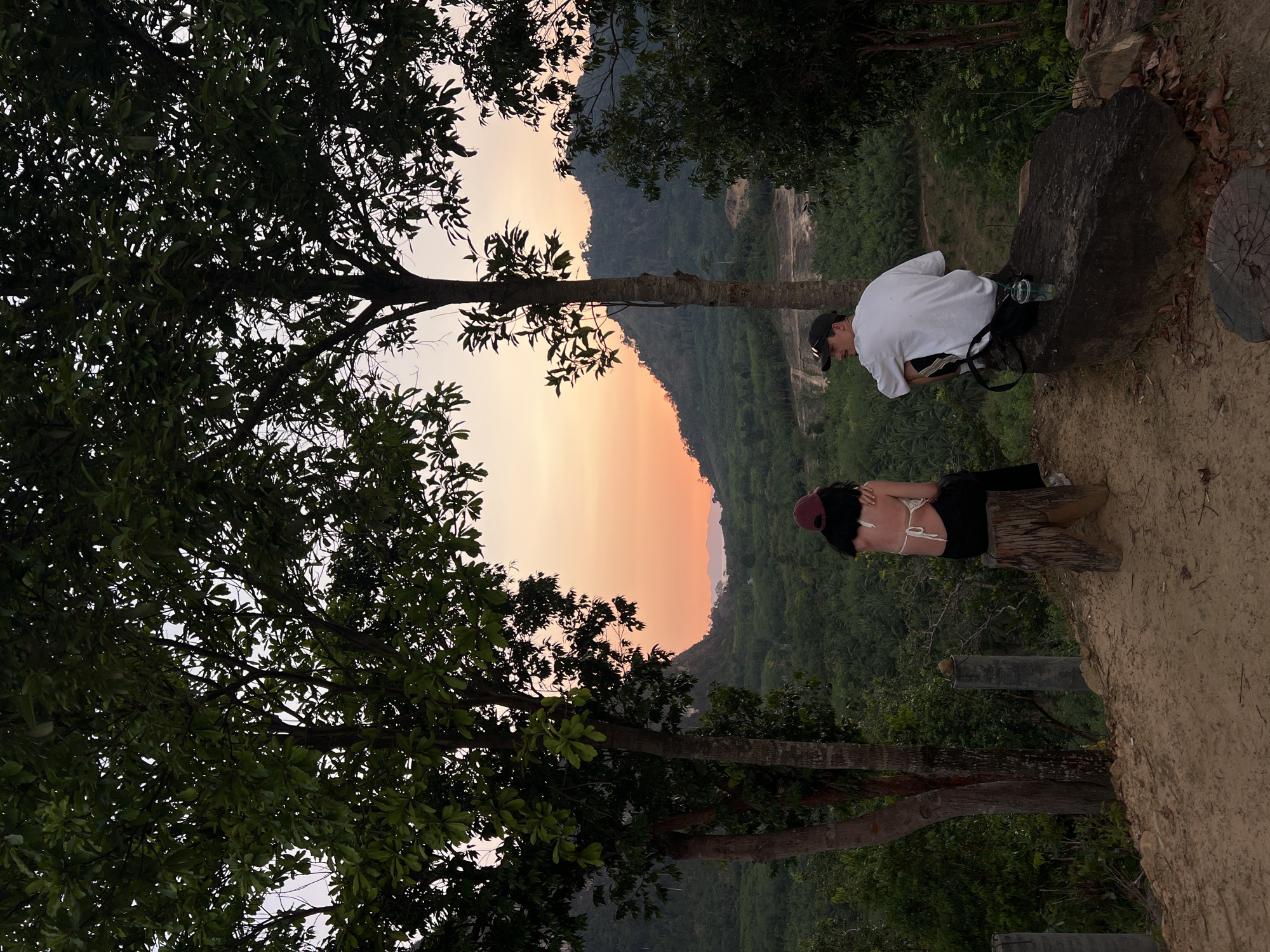 Two people are positioned on the edge of a ridge overlooking a green, hilly landscape near Khao Thong, Thailand, as the sun sets, casting a warm glow behind them. A man wearing a white t-shirt and a black cap is seated on a large rock, while a woman wearing a maroon cap and a white string top sits on a wooden log. The foreground features silhouetted tree branches that partially obscure the view.