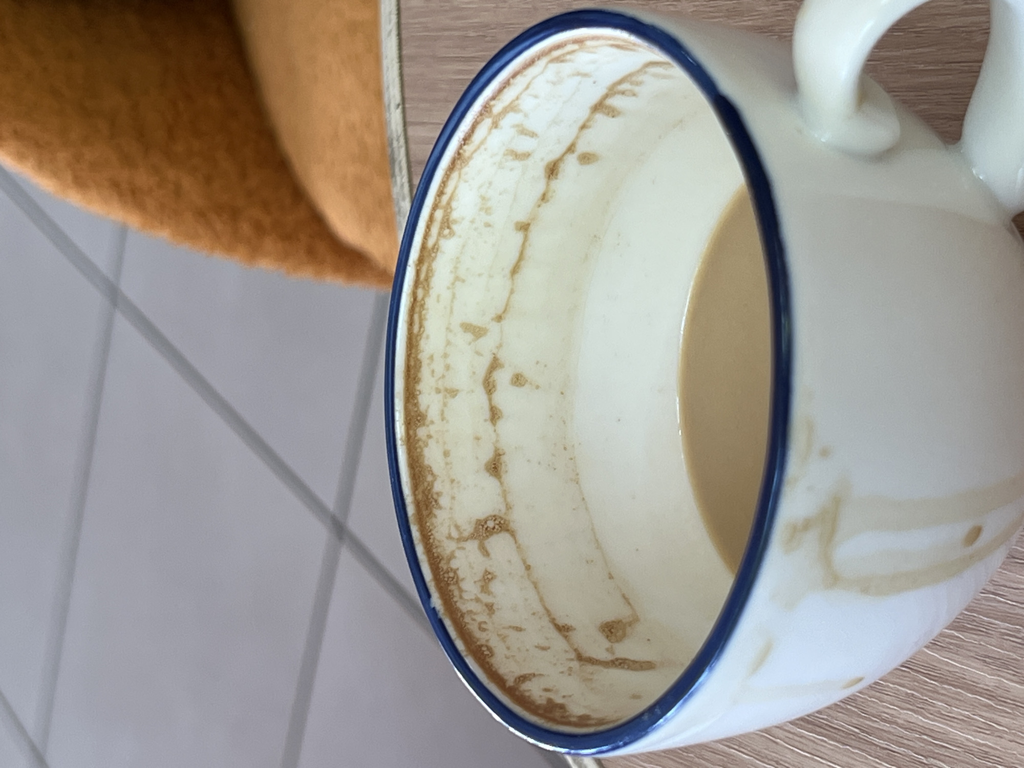 An extreme close-up, eye-level shot shows the interior of a white ceramic coffee mug with a blue rim. The mug contains a small amount of light brown liquid, possibly coffee with milk. Streaks of dried coffee residue are visible along the inner rim and dripping down the interior walls of the mug. The exterior of the mug also shows a few dried drips. The mug rests on a light brown wooden surface with a visible grain pattern. In the blurred background, a portion of a textured, orange chair is visible, as is a section of a pale gray tiled floor with intersecting dark gray grout lines. The scene suggests the interior of a cafe or a home in Antalya, Turkey.