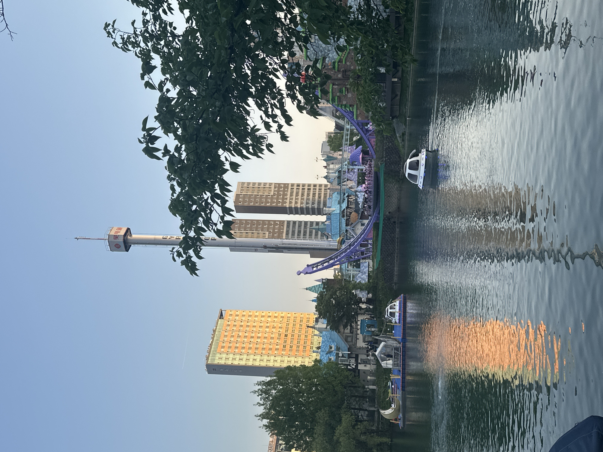 The image captures a scenic view in Seoul, South Korea, featuring a lake or river reflecting sunlight. In the background, there is a prominent amusement park with a tall observation tower, likely part of Lotte World, a major recreation complex in Seoul. There are also roller coasters and themed structures. Several high-rise buildings are visible in the background, contrasting with the leisure-focused foreground. The presence of watercraft on the water indicates recreational activities. The image is taken during twilight or golden hour, with the sunset casting a warm glow on the buildings and water. Trees frame the scene, adding a natural element to the urban landscape.