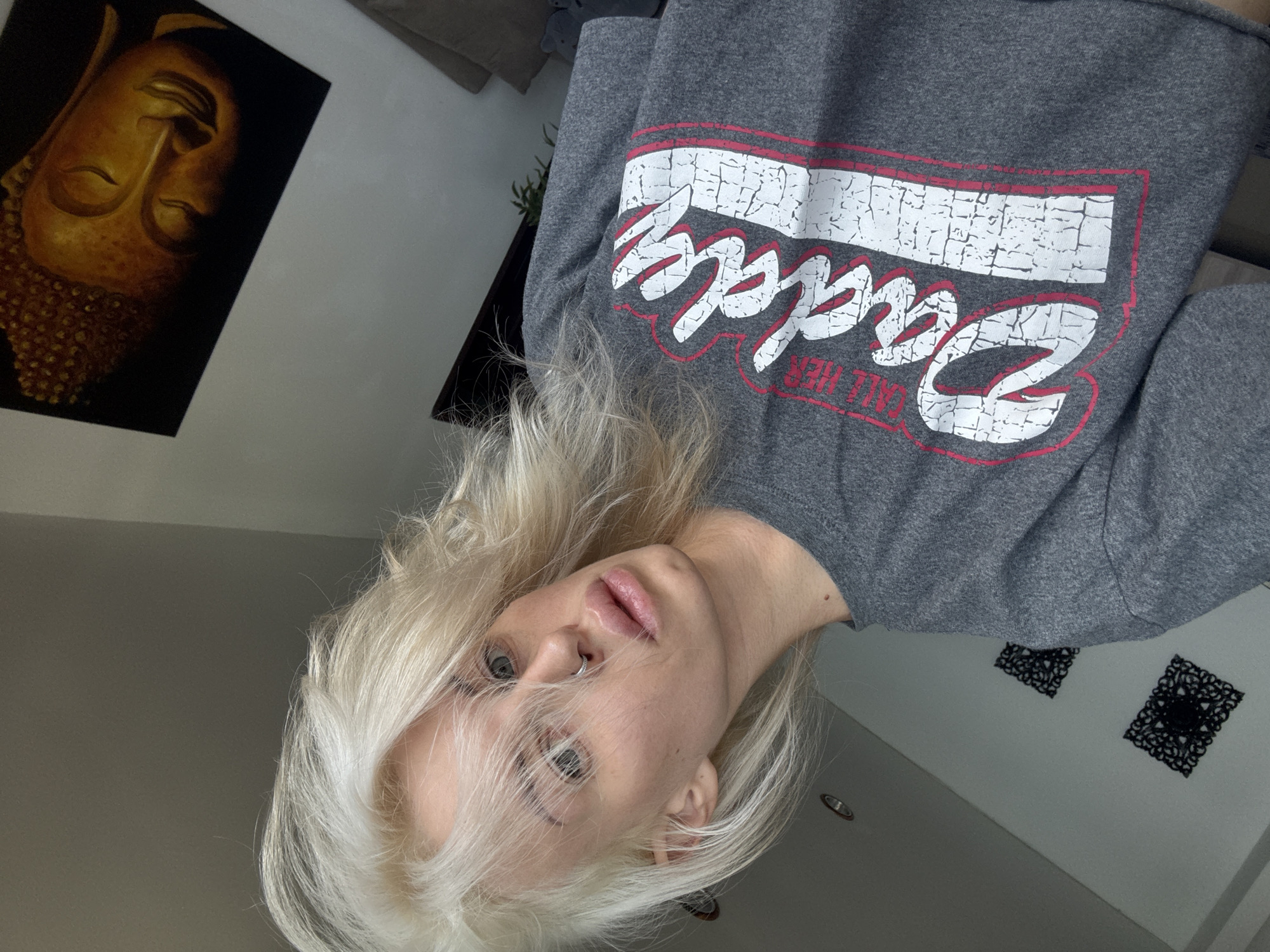 The image is a selfie taken indoors. A woman with platinum blonde hair, a nose ring, and fair skin is in the foreground. She is wearing a gray sweatshirt with "Daddy" printed on it in white letters with a red outline. Behind her, a wall is decorated with a black and gold Buddha painting, and two black, patterned decorations. The environment suggests a modern, possibly urban interior. The location in Baan Chaweng Noi, Thailand is speculative, based on the presence of Buddhist imagery.