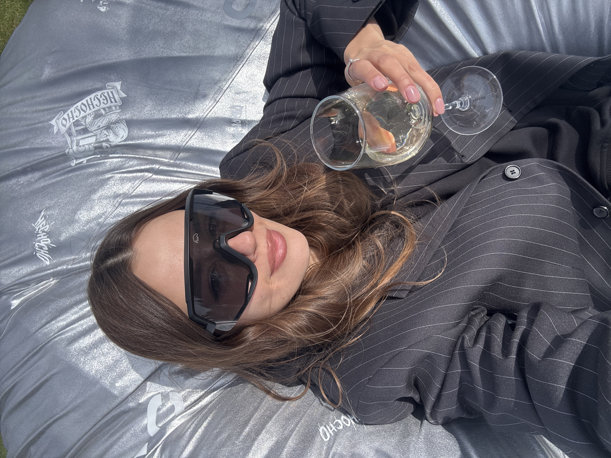 A young woman in a dark pinstripe suit and large sunglasses is lying down, holding a glass of light-colored liquid, possibly white wine. She is on a silver-colored surface with a graphic design. The location is likely outdoors given the presence of natural light. The city of Krasnoyarsk is not discernible from the image.