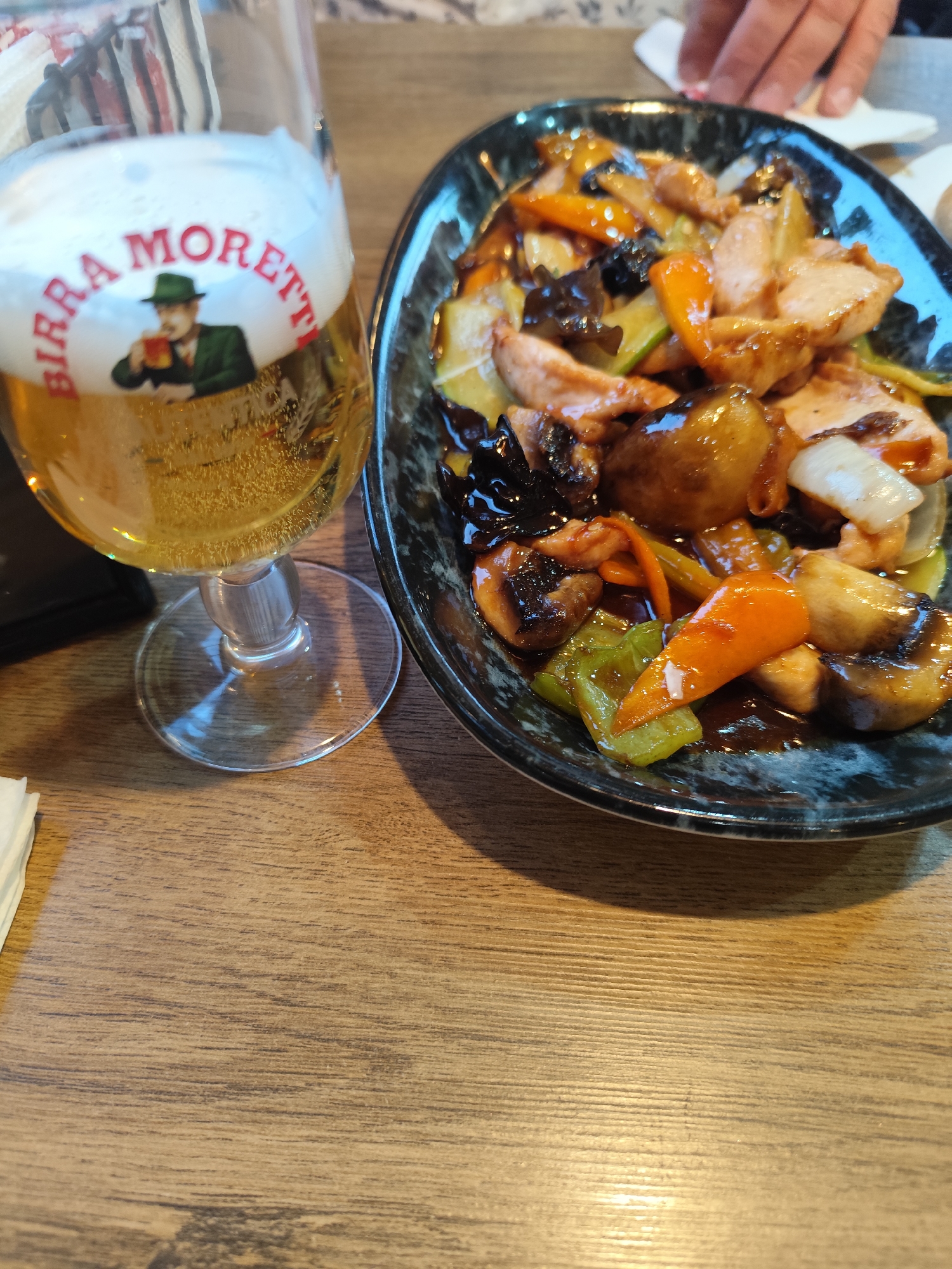 A close-up shot shows a glass of beer with the logo "BIRRA MORETTI" visible on it, alongside a plate of stir-fried food containing chicken, vegetables, and mushrooms. The food is coated in a dark sauce and served in a patterned bowl. The scene is set on a wooden table. The image does not provide information about its location in Montana, Bulgaria.
