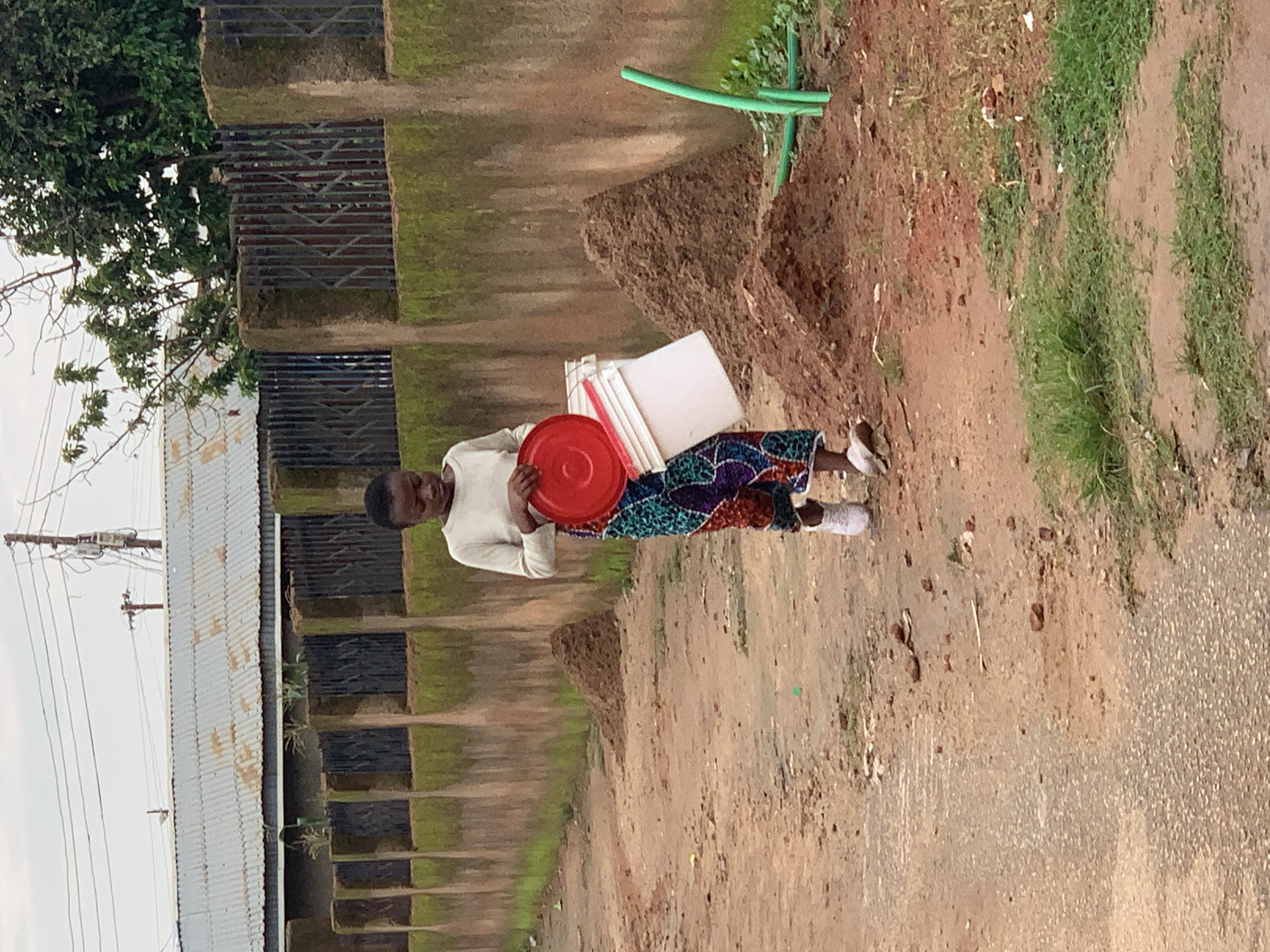 A young person, wearing a light-colored long-sleeved shirt and a vibrantly patterned skirt, walks on a dirt path in a residential area of Jos, Nigeria. The person is carrying a red lid in their left hand and a white bucket with a red handle in their right hand. The ground is a mix of dirt, loose soil, and sparse green vegetation. In the background, a moss-covered concrete wall with a metal grate separates the path from a property with a corrugated metal roofed building. Several mounds of soil are visible along the side of the path. A green plastic pipe snakes across the ground near one of the soil mounds. The sky is overcast.