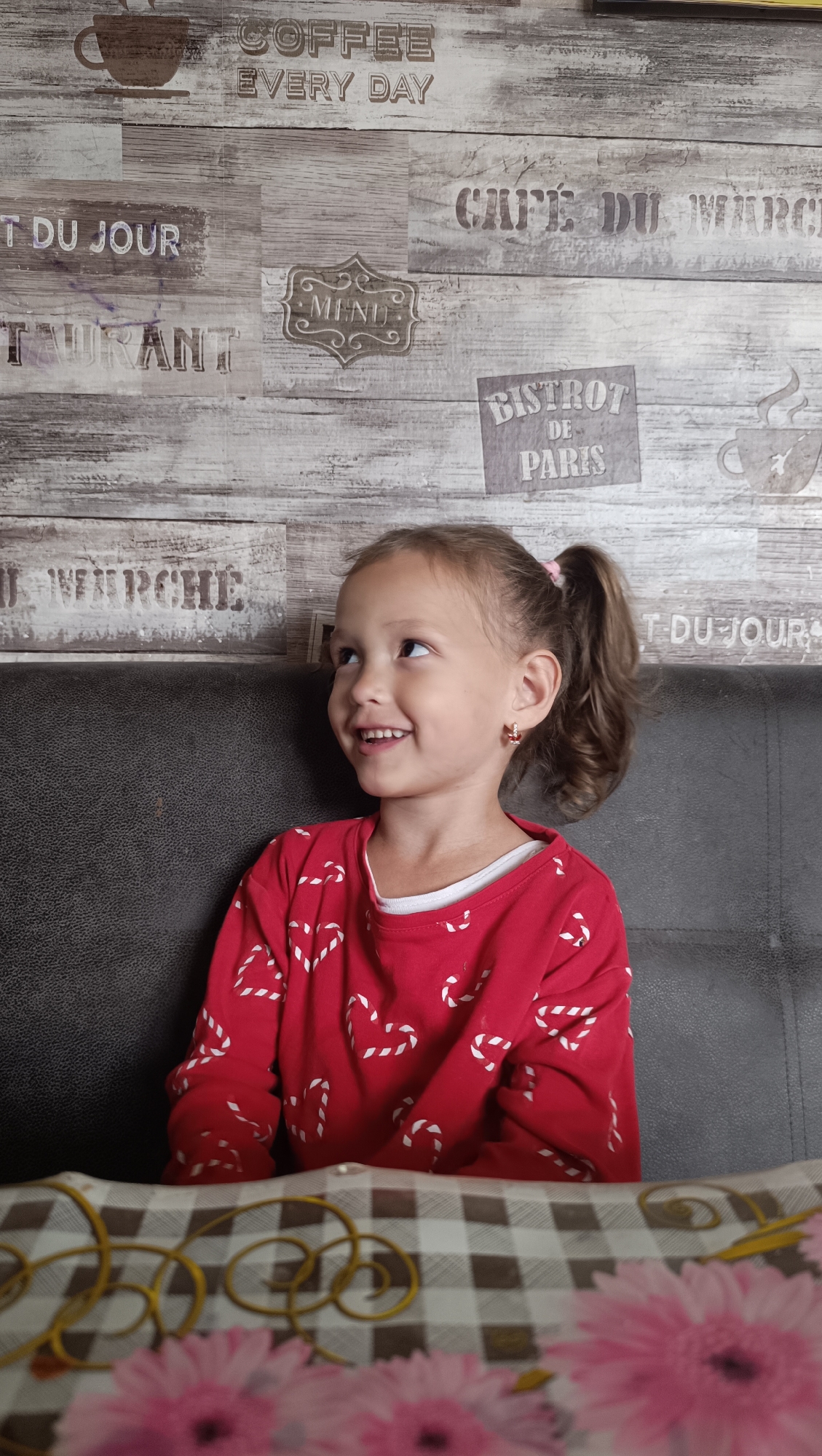 The image depicts a young girl with a ponytail, wearing a red long-sleeved shirt adorned with white candy cane heart patterns, sitting on a dark leather couch. She is looking to her left and smiling. The background features a wall with a wood-like texture and vintage cafe-themed text, including "COFFEE EVERY DAY," "CAFE DU MARCHE," "MENU," "BISTROT DE PARIS," and "T DU JOUR." In the foreground, a table is partially visible, covered with a checkered tablecloth and decorated with yellow curly strands and pink flowers. The location is specified as the city of Karsha, Russia.