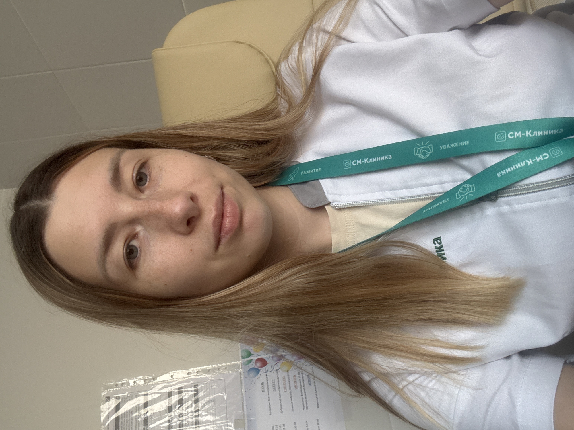 The image shows a woman with light brown hair, wearing a white lab coat and a green lanyard with "CM-Клиника" (SM-Clinic) printed on it in Russian. The lanyard also has the words "РАЗВИТИЕ" (Development) and "УВАЖЕНИЕ" (Respect) written on it. In the foreground, there is a document with some Russian text visible, including the name "Муратова Пасьяна Никологана" and dates like "13.10". The background includes a beige chair and a white wall. Based on the "CM-Клиника" on the lanyard, the location is likely related to the SM-Clinic network, which has multiple locations in Moscow, Russia.