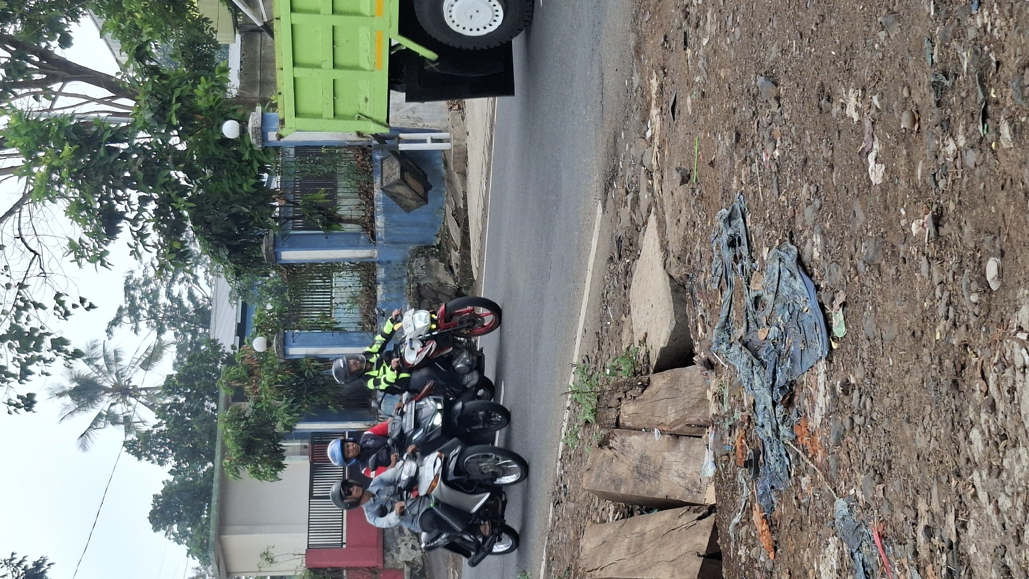 In Cilaku, Indonesia, three individuals are riding motorcycles on a paved road. The foremost motorcycle, silver, carries two individuals wearing helmets. The second motorcycle, black, has one rider wearing a red jacket and blue helmet. The third motorcycle, red, has a rider in a neon yellow and black jacket and helmet. To the right, a large green dump truck is partially visible, with its rear wheel and portion of the bed facing the viewer. The environment includes a blue and white fenced area with buildings and trees in the background. The foreground shows a dirt area with scattered debris, including pieces of wood, fabric, and rocks. The sky is overcast.