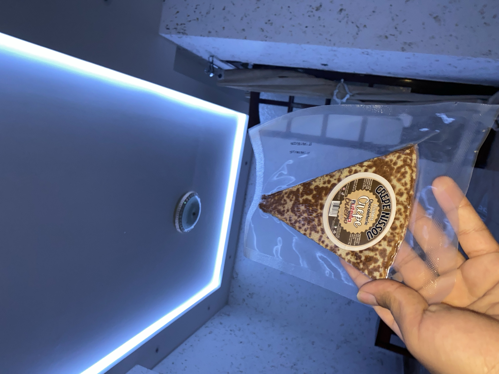 A hand is holding a triangular-shaped crepe, likely a dessert, sealed in clear plastic packaging. The crepe appears to have a chocolate-based filling or topping, visible as a speckled pattern. A circular label is affixed to the crepe, with text that includes "CREPE," "CHOCOLATERIE," and "NISSU." The background shows a modern ceiling with blue LED lighting, suggesting an indoor setting. To the right, a portion of a wall with a textured finish is visible. The overall lighting suggests the photo was taken at night or in a dimly lit interior, illuminated by the artificial lights. The presence of the crepe and the modern interior design does not provide specific locational information within the city of Baraki, Algeria, beyond it being an indoor location.