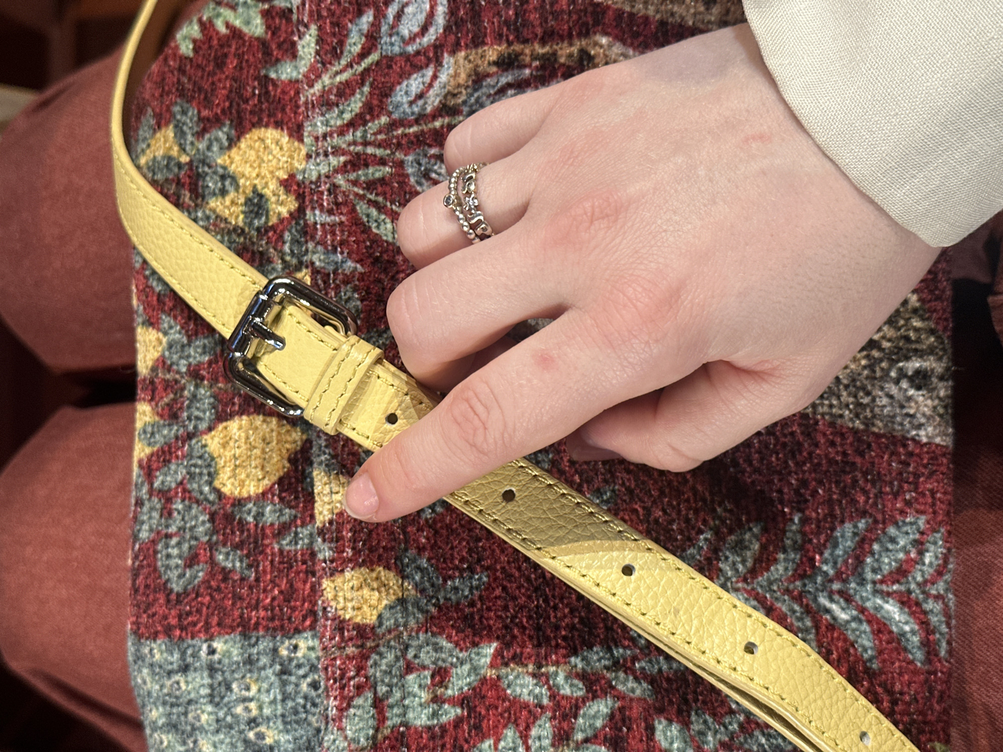 A close-up shot captures a person's hand interacting with a yellow, textured strap. The hand, with fair skin and neatly trimmed nails, wears a multi-banded ring on the ring finger, featuring small embellishments or stones on silver-toned metal. The index finger points towards one of the holes in the strap, suggesting an action of adjusting or examining it. The strap itself has a shiny silver buckle.
The strap rests upon a richly patterned fabric that dominates the background, displaying an intricate, possibly floral or abstract, design in hues of deep red, mustard yellow, and muted blue-grey. To the left, a plain reddish-brown fabric provides an additional background element. A white or off-white fabric cuff is visible on the person's wrist, near the top right of the frame. The image does not contain any discernible text, and no specific time of day or emotions can be inferred. The scene is situated in Moscow, Russia.