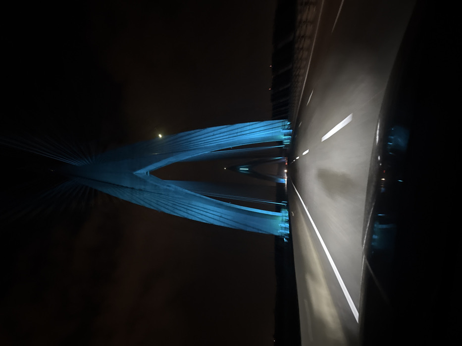 A nighttime view from a vehicle moving on a highway. A large, blue-lit bridge towers beside the road, its supporting cables fanning out into the dark sky. The road stretches into the distance, illuminated by vehicle headlights. Lanes are clearly marked. A car's side mirror is visible in the right foreground, reflecting the passing scene. The sky is dark with hints of mist or low clouds.