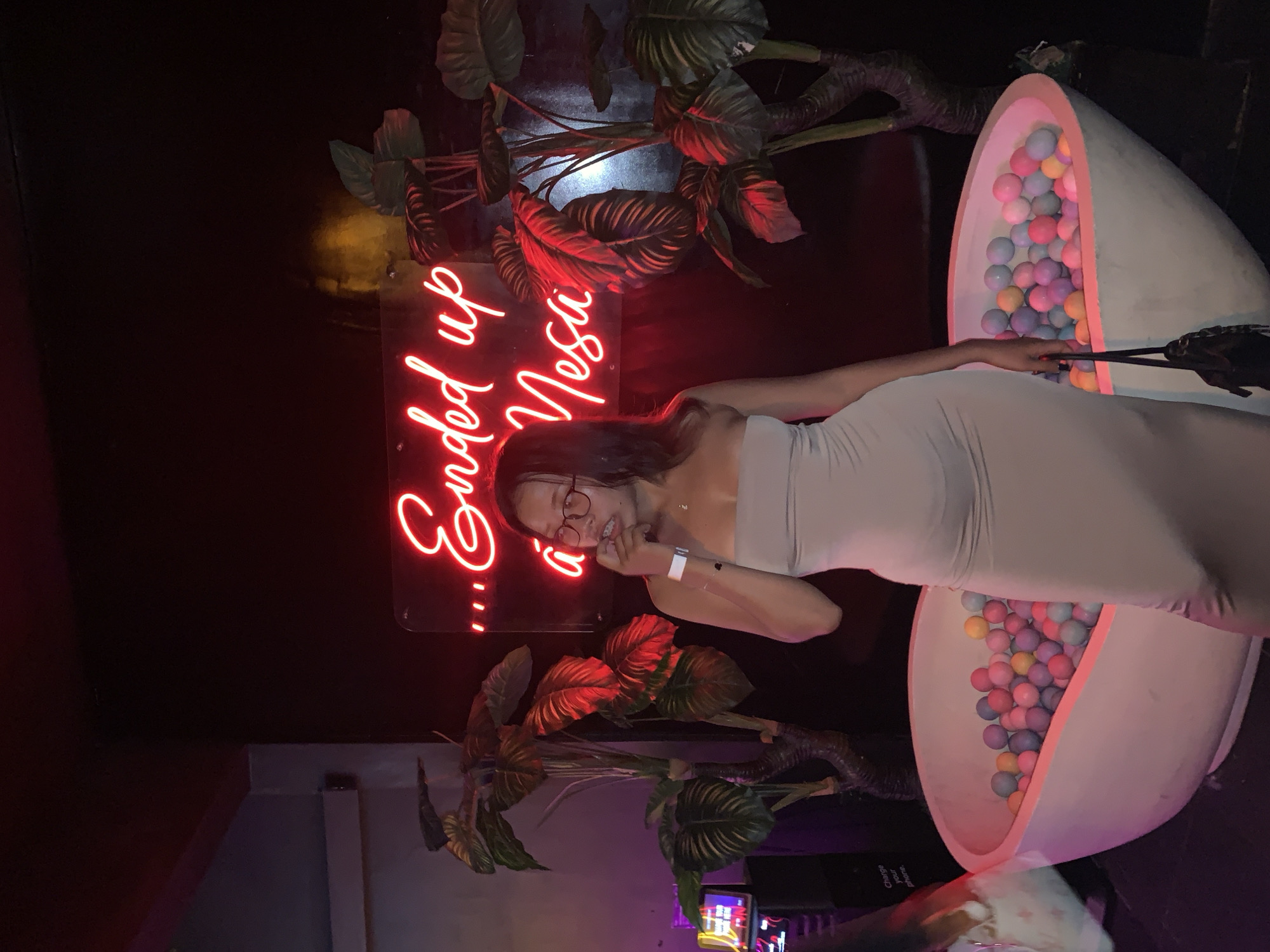 A woman wearing a light beige dress is posing in front of a neon sign that reads "...Ended up Nesú" and decorative plants. She is also near a large egg-shaped structure filled with colorful balls. The location is identified as North Kuta, Indonesia. The woman has glasses and a watch on her wrist. She is holding a black purse with straps.