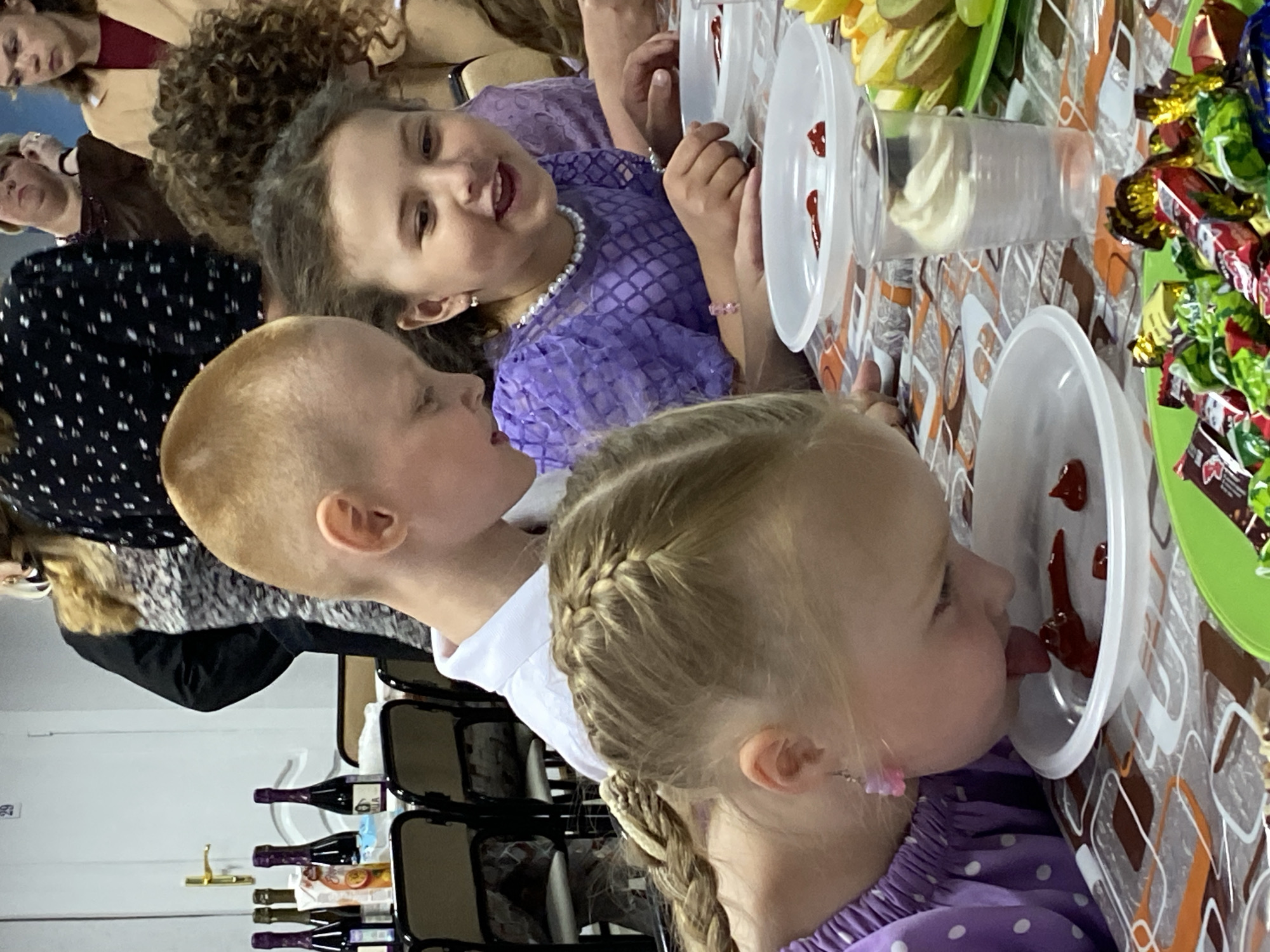The image captures a gathering, potentially a party or event, in what appears to be an indoor setting. Several individuals are present. Three young children are the most prominent subjects. One child with short blonde hair looks towards another child with a braided hairstyle who is shown licking a substance from a white plate. The third child, a girl in a purple dress and a pearl necklace, is looking at the other two. A table is visible, laden with various items including plates with a reddish substance, cups filled with what appears to be whipped cream, fruits (kiwi), and wrapped candies. In the background, several adults are present. Additionally, chairs and what seems to be a collection of wine bottles are observable near a white wall. There is no information to confirm the location as Noviy, Russia.
