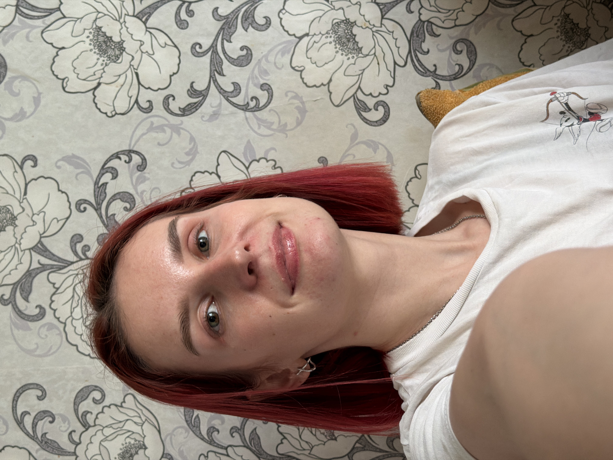 A young woman with red hair is taking a selfie indoors. She is wearing a white t-shirt and a silver necklace. She has fair skin, green eyes, and light eyebrows. The background is a wallpaper with a floral pattern in gray and white. The location is Заболотово, Russia.