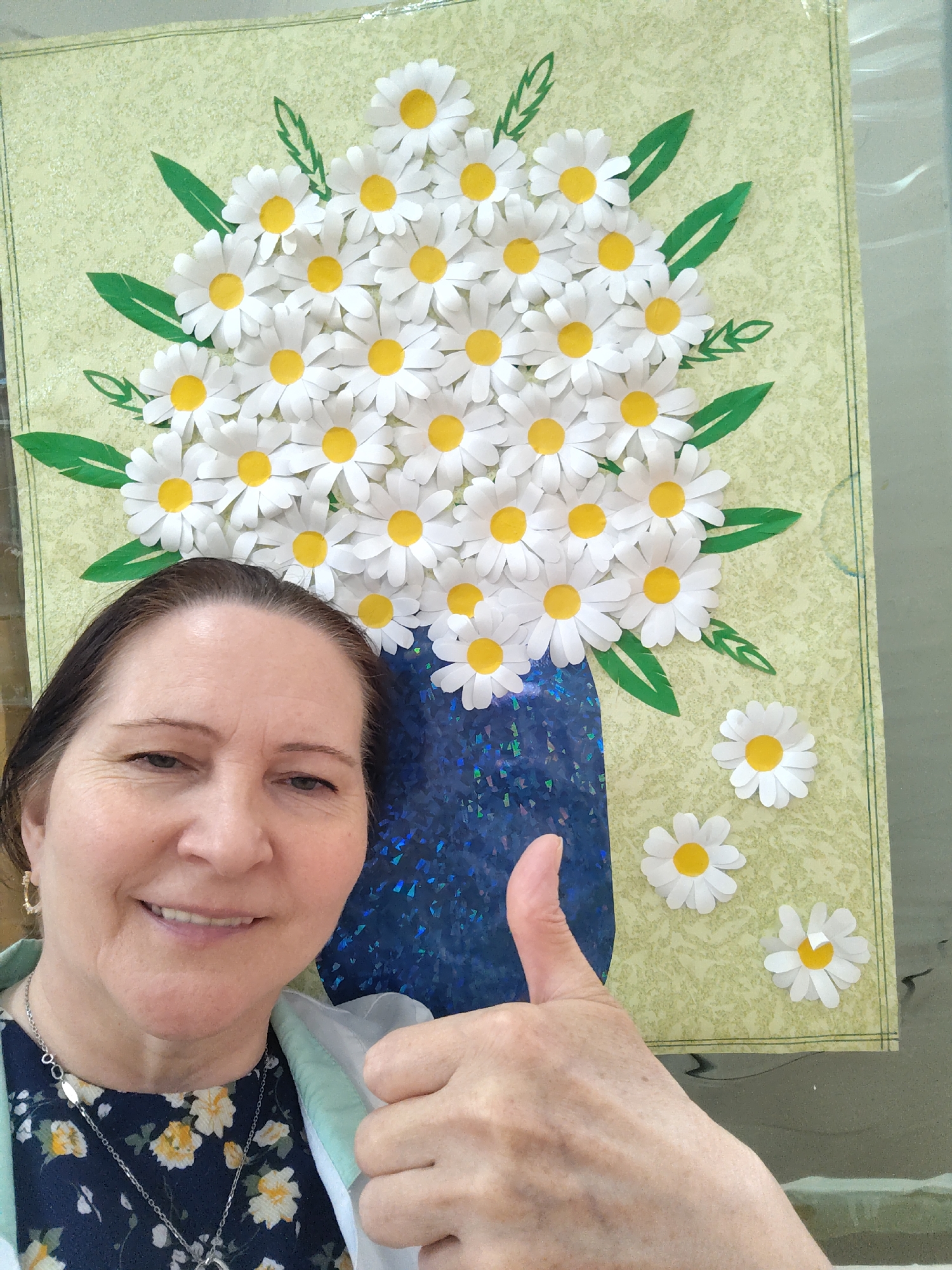 A person is holding their thumb up in front of a framed artwork depicting a vase of daisies on a light green background. The daisies are white with yellow centers and are arranged in a bouquet shape, with green leaf accents. The vase is blue and iridescent. The person is wearing a navy blue floral shirt, a light green jacket, and jewelry. The environment appears to be indoors. There is no information in the image to determine the location in Sarapul, Russia.