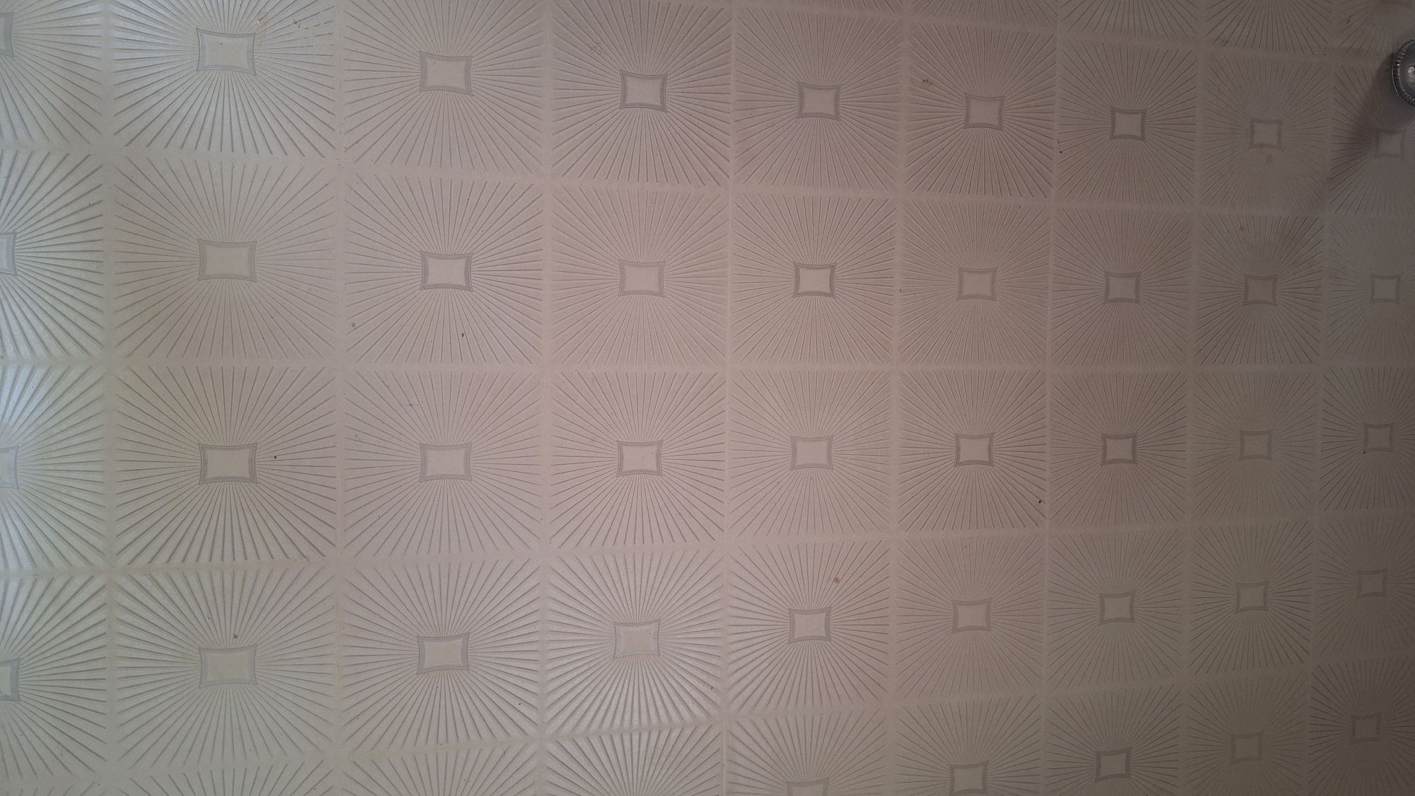 The image shows a close-up, slightly angled view of a textured ceiling. The ceiling is tiled with square panels, each featuring a decorative pattern that resembles radiating lines emanating from a central square. The pattern is repeated across the entire visible surface of the ceiling. The lighting appears to be coming from above and slightly to the right, casting subtle shadows that accentuate the texture. The overall color palette is neutral, with variations in shading due to the light and shadow. No people or specific objects are clearly identifiable within the frame, beyond the architectural feature of the ceiling itself. The location is unknown, as is the specific context of the image.