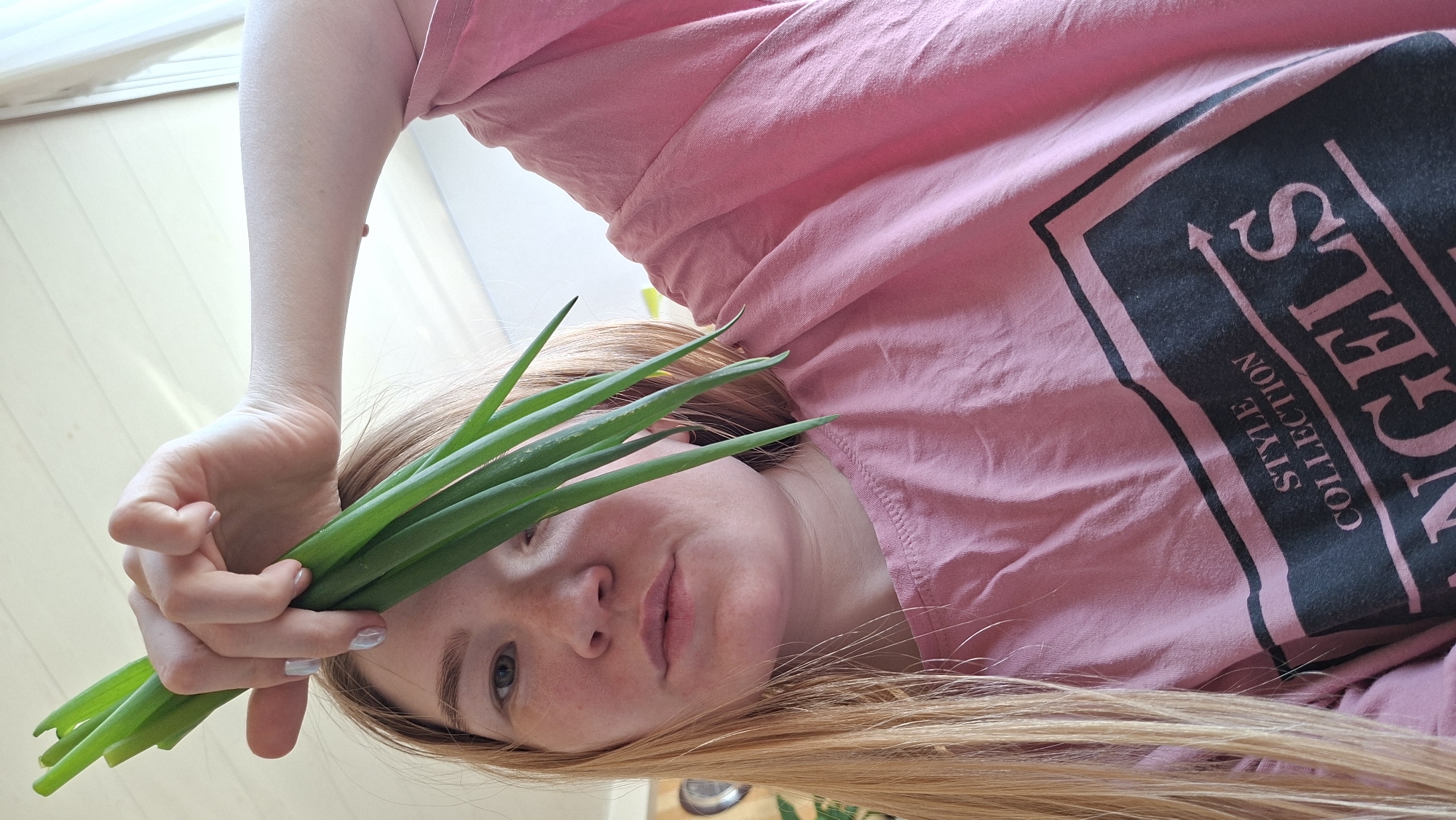 A fair-skinned woman with light blonde hair is holding a bunch of green onions in front of her face. She's wearing a pink t-shirt with a black rectangular design containing lettering, partially obscured but includes the word "ANGELS" and "STYLE COLLECTION." Her left hand is visible, holding the onions. The background appears to be an interior setting with a wooden floor. Location: Большой Букор, Russia.