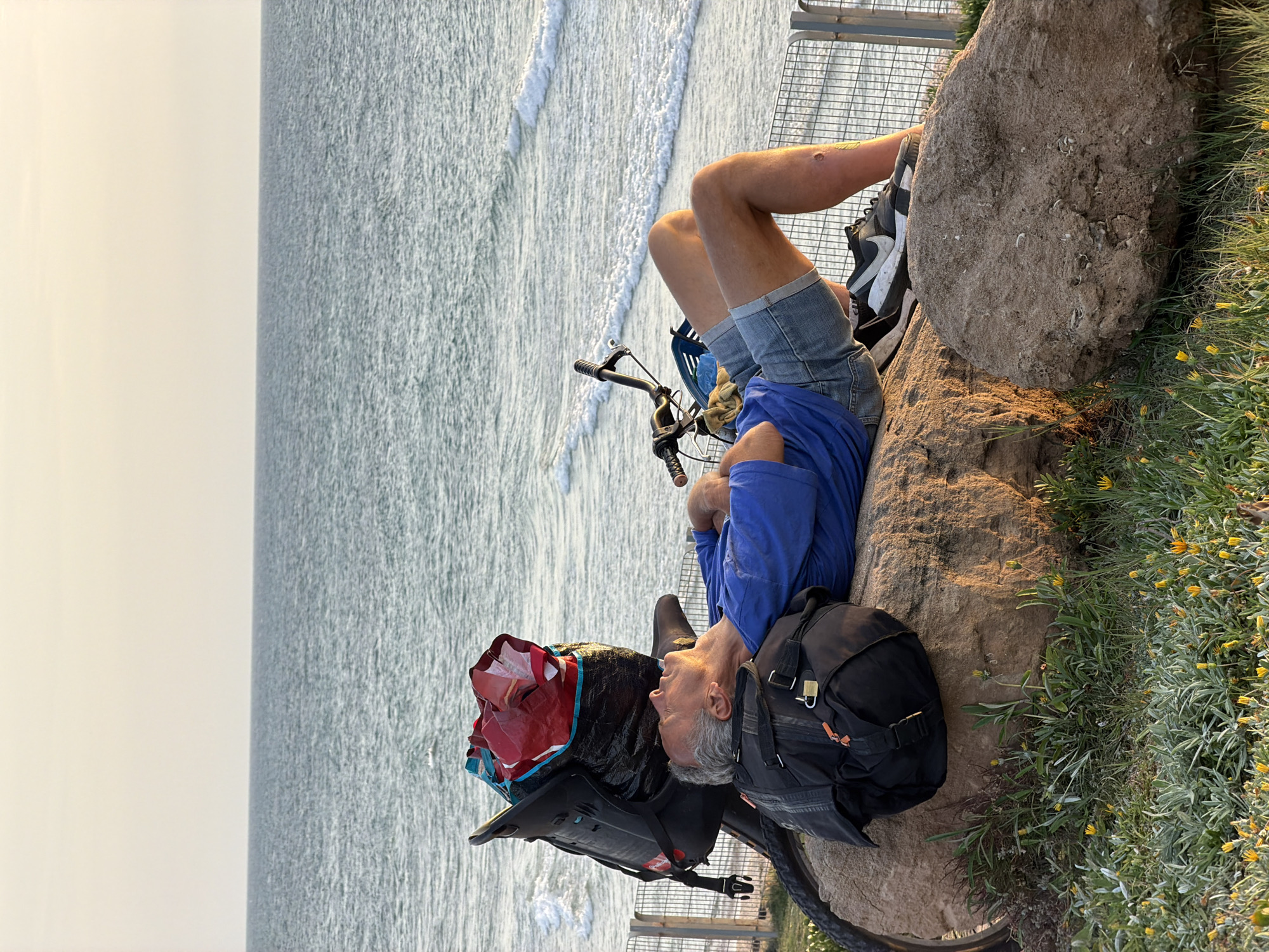 A silver-haired man wearing a blue shirt and jean shorts is lying on a rock next to his bicycle, overlooking the Mediterranean Sea. The bicycle is equipped with panniers and bags, suggesting a long journey or touring. The sea is calm with gentle waves breaking on the shore. The scene is located near a metal fence with vegetation and yellow flowers growing adjacent to the rock. The location appears to be along the coastal path in Tel Aviv, Israel.