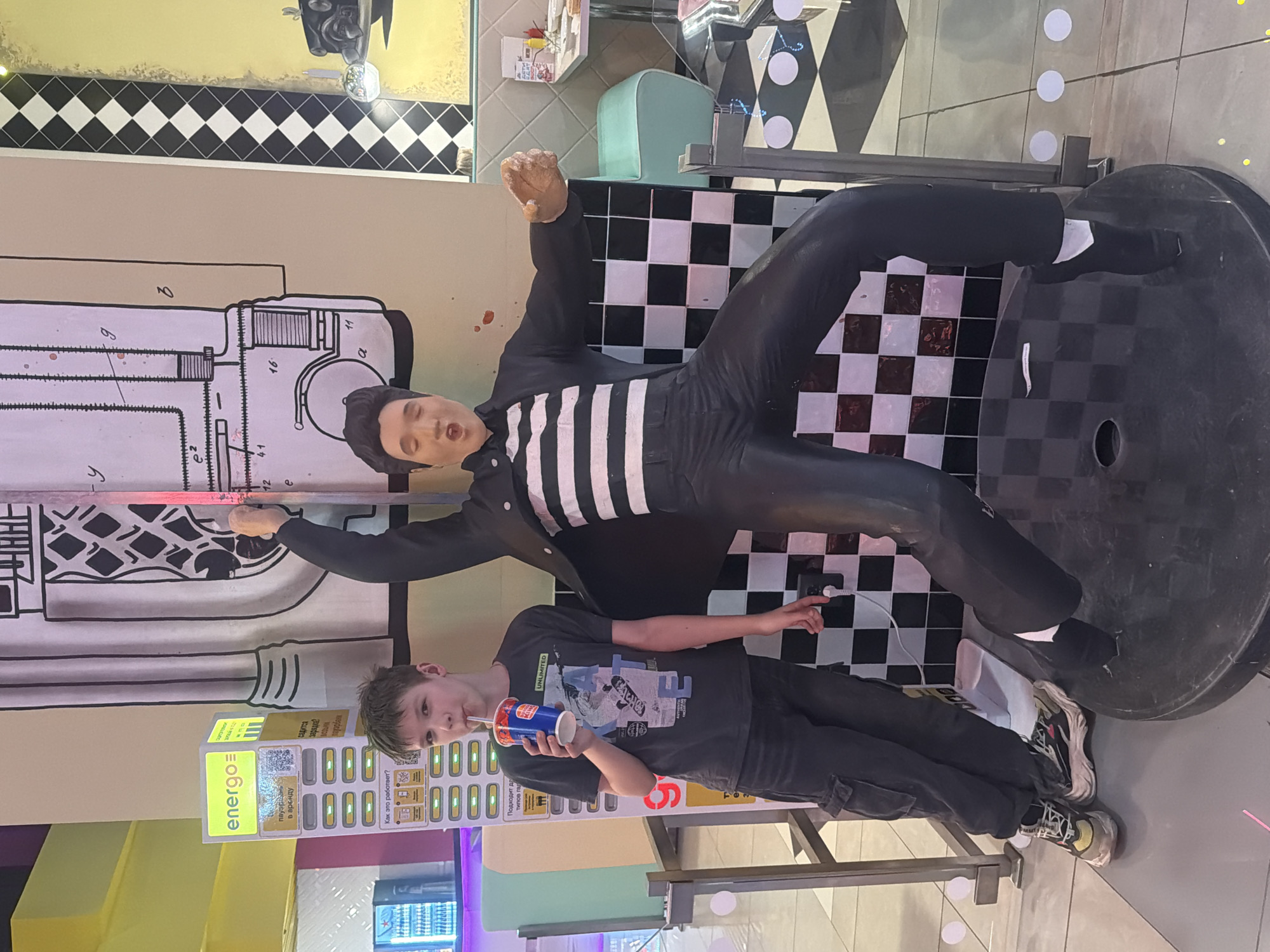 The image displays an indoor scene within a retro-themed establishment, identified as a food court or entertainment venue in Perm, Russia, due to the presence of Russian text on a charging station. A male individual, appearing to be a child or young teenager, stands on the left, looking forward while drinking from a Burger King cup with a straw. He is dressed in a dark grey graphic t-shirt featuring a skateboarder motif and the word "UNLIMITED," dark cargo pants, and multi-colored athletic sneakers. His left arm rests on a silver metal barrier. Immediately to his right, a life-sized, full-body statue of Elvis Presley is depicted in a dynamic, mid-action pose, clad in a black jacket, a black and white striped shirt, and black pants, standing on a round, black and white checkered base. Behind the boy, a prominent yellow and white "energo=" charging station is visible, featuring multiple charging compartments and Russian text, including phrases like "подходит для типов гаджетов" (suitable for types of gadgets) and "Как это работает?" (How it works?). The background wall behind the statue is light beige, adorned with a large black and white diagrammatic illustration. This transitions to a vertical black and white checkered strip, followed by a yellow wall section exhibiting a partial illustration of a vintage vehicle. In the far right background, light teal diner-style booth seating and a white table holding condiments are visible. The floor is a light grey surface, sporadically marked with white circular patterns. A commercial beverage cooler containing various drinks is partially visible in the far left background.
