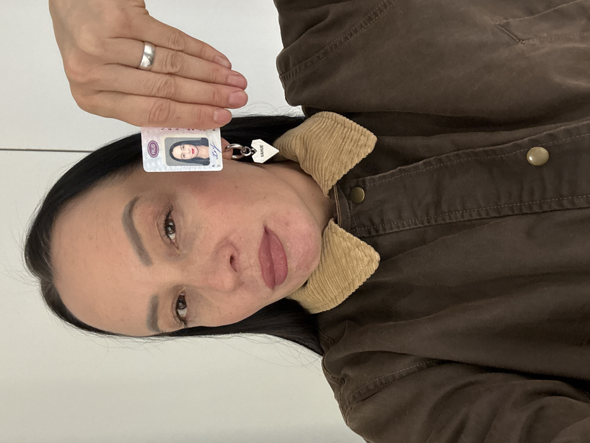 This image depicts a woman in an indoor setting in Chaikovsky, Russia. The woman, with dark hair and dark eyes, wears reddish-pink lipstick and a dark brown jacket featuring a light brown corduroy-style collar and brass-colored buttons. She is looking directly forward. Her right hand is positioned near her face, holding a small identification card vertically. The card displays a photograph of a woman resembling the subject, the abbreviation "RUS" at the top, and partially visible Cyrillic script. A silver-colored band ring is present on her left ring finger. A silver-colored hoop earring, from which a heart-shaped pendant engraved with "VAHUE" hangs, is visible in her left ear. The background consists of a plain, light-colored wall.