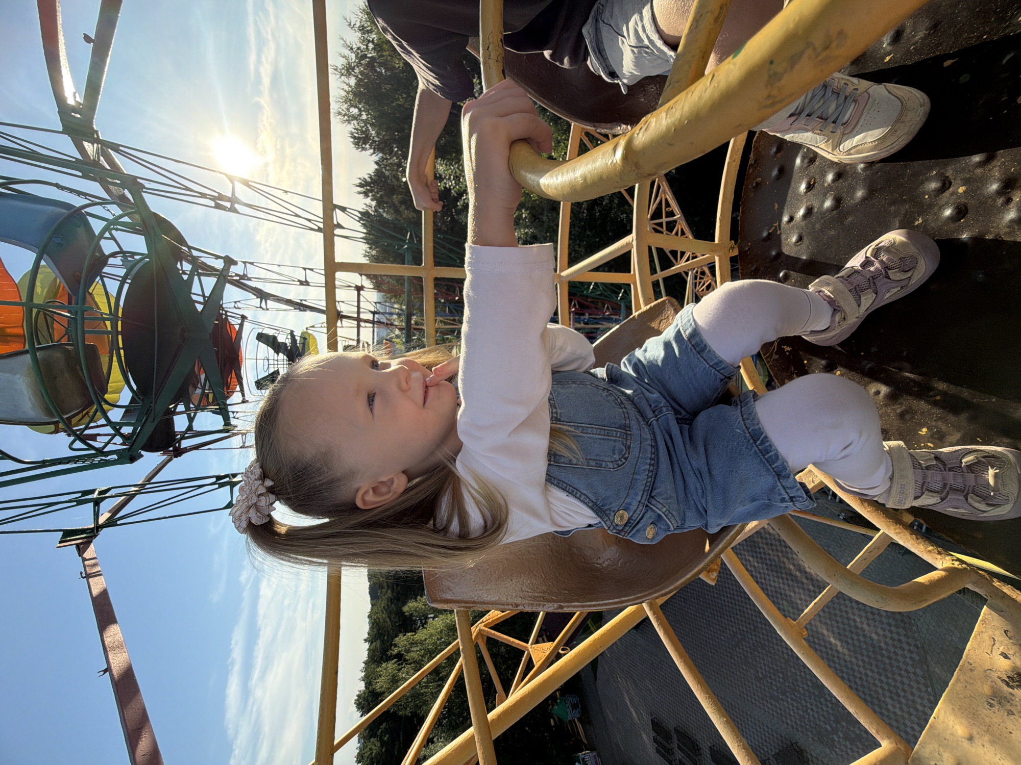 A young girl with blonde hair tied in a ponytail with a scrunchie is sitting in a gondola of a Ferris wheel, likely at an amusement park in Chaikovsky, Russia. She is wearing a white long-sleeved shirt and denim overalls. Her right arm is resting on a yellow metal bar of the gondola. The sun is shining brightly in the sky, casting a lens flare. In the background, the structure of the Ferris wheel and trees are visible. Another person's legs are visible to the right of the girl. The ground below appears to be paved.