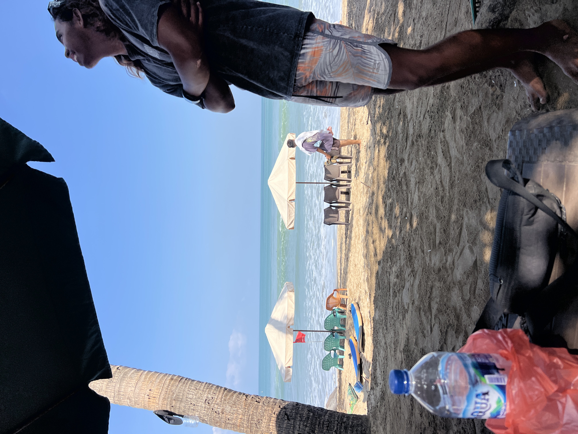 A man with long, curly hair and arms crossed is looking up at the sky on a sunny day at Kuta Beach, Indonesia. He is wearing a dark t-shirt and patterned shorts. In the foreground, to the left, is a red plastic bag with a bottle of Aqua water. Also in the foreground is a black bag. The beach is sandy, and further in the background are two beach umbrellas, several chairs, and two surfboards. The ocean is visible beyond the beach, with gentle waves. A person in a purple top and dark shorts is walking away from the camera near one of the umbrellas.