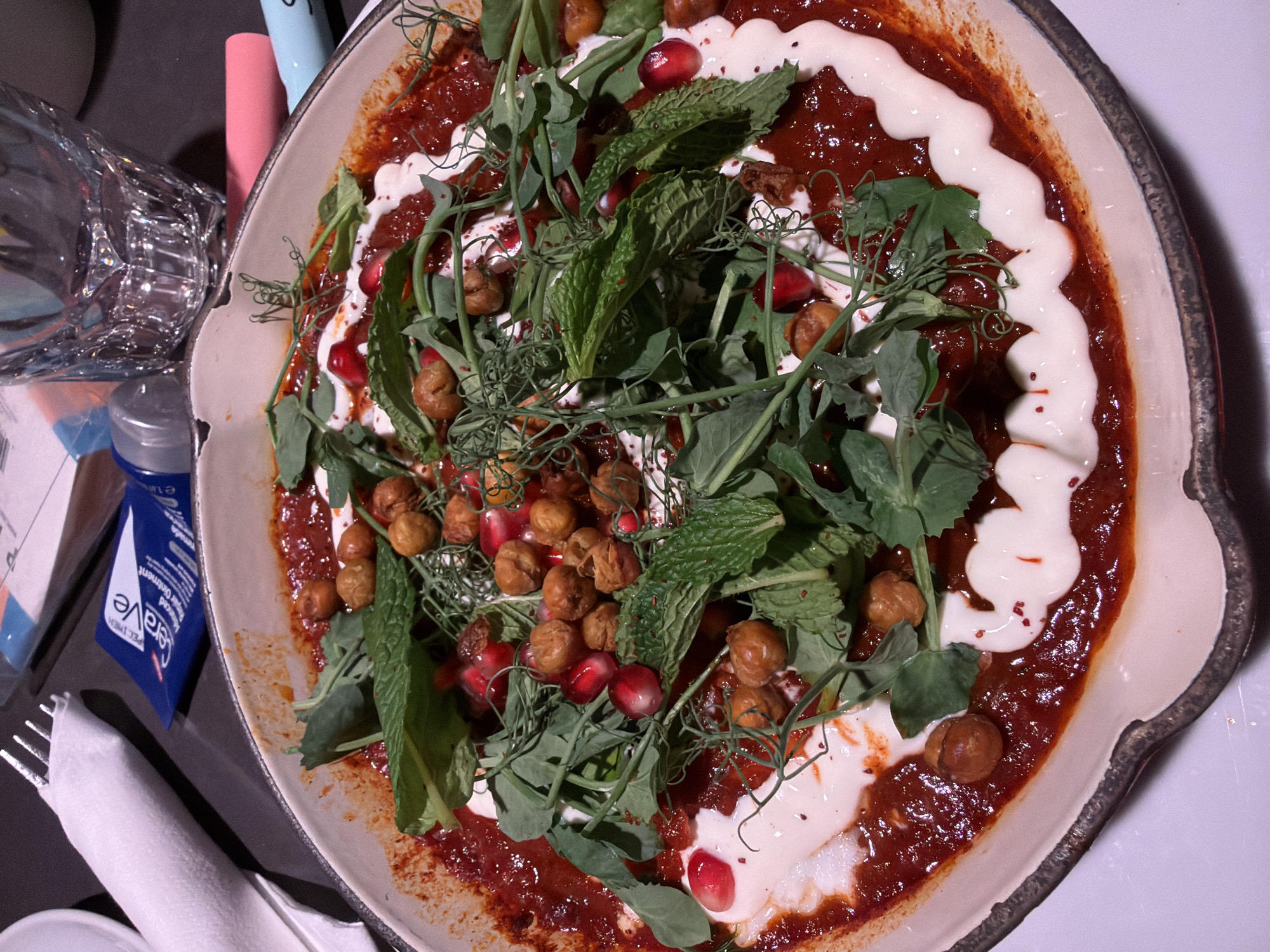 The image depicts a close-up of a dish served in a shallow, ornate bowl. The dish consists of a reddish-brown sauce, possibly tomato-based, arranged in a ring. A row of white sauce is placed on the base along the inner rim of the plate. The sauce is topped with chickpeas, fresh mint leaves, pea shoots, and pomegranate seeds. A fork, a napkin, tubes of a cream-colored lotion and a blue ointment, and a clear glass are visible on the left side. The scene appears to be a restaurant setting, potentially on a dining table. There is no explicit evidence indicating that this image originates from Montreal, Canada, but no information exists to disprove it.