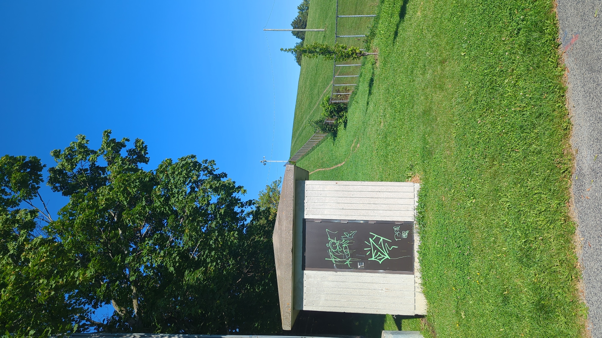 This is an eye-level, outdoor, full-shot photograph taken on a clear, sunny day. The scene depicts a small, rectangular concrete utility building with a sloped, light-colored roof, situated on the left side of the frame. The building's front is clad in vertical, pale gray siding, and the dark metal door is covered in green graffiti.
To the right of the building, a gently sloping, vibrant green grassy hill rises towards a clear, bright blue sky. A metal chain-link fence runs diagonally up the hill, following a path. Several small trees and shrubs are visible on the slope, along with faint tracks in the grass, suggesting a well-trodden path. A tall utility pole with lights is visible near the crest of the hill, and another pole is further to the right. The foreground consists of a patch of well-maintained green grass leading to a gray asphalt path. The background shows more trees and the clear blue sky. The overall impression is one of a park or open recreational area in a suburban or semi-rural setting within Montreal, Canada.