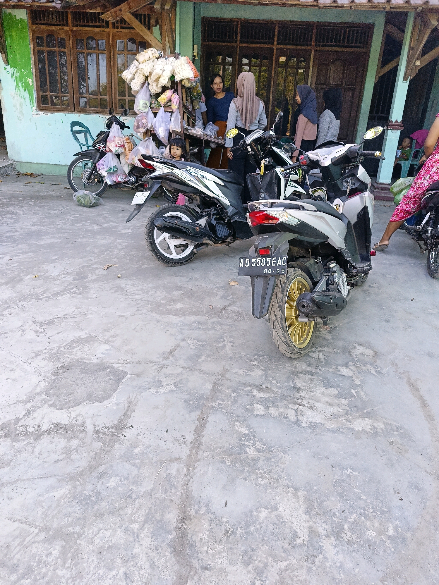 This is a horizontal, medium-shot photograph taken from a low angle, depicting a street scene in Sukorini, Indonesia. The foreground is dominated by a grey concrete ground, which shows signs of wear and tear with visible cracks and patches of lighter and darker tones.
In the middle ground, several motorcycles are parked. The most prominent is a silver and black scooter on the right, facing away from the camera. It has a license plate with the registration number "AD 5505 EAC" and a date "08.25" below it. The scooter's rear wheel features a distinctive gold rim. To the left of this scooter is a black motorcycle, also facing away, loaded with several plastic bags of snacks or goods attached to its sides and handlebars. A young child with dark hair is seated on this motorcycle, peeking out from behind the bags.
In the background, a light blue building with wooden window frames and a tiled roof stands. In front of the building, a stall is set up, displaying numerous plastic bags filled with white snacks. Several individuals are gathered around this stall. From left to right, a woman with dark hair and a blue top is visible, followed by three women wearing headscarves. One woman is wearing a beige headscarf, another a blue one, and the third a grey one. They appear to be interacting or observing the goods. Further to the right, a person in a pink and white patterned garment is seated on a motorcycle, partially visible. To the far right, another person in a green shirt is seated on a blue plastic chair. The overall atmosphere suggests a local market or a roadside shop.