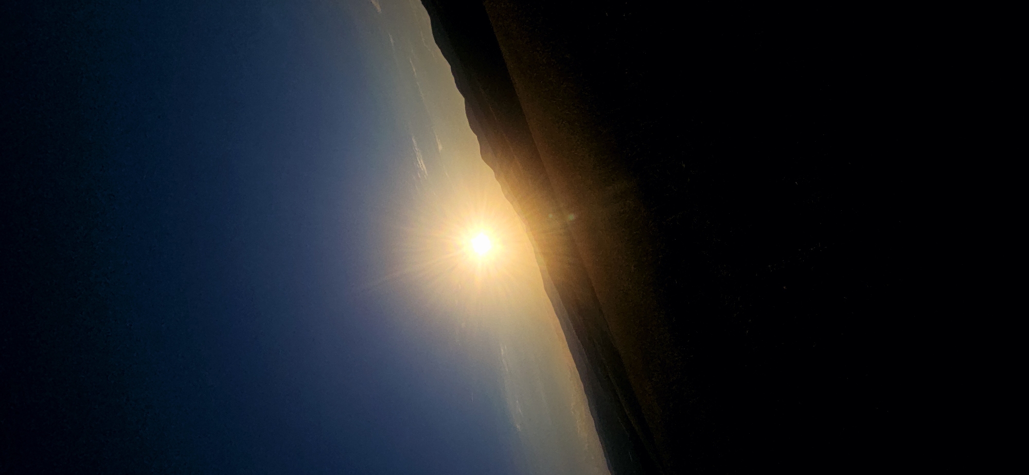 The image captures a striking sunset over a landscape of rolling hills. The sun, a brilliant orb of light, is positioned in the upper middle of the frame, casting a warm golden glow across the scene. Rays of sunlight emanate outwards, creating a starburst effect. The sky transitions from a deep, dark blue at the top of the image to a lighter, softer blue near the horizon, with subtle hints of white clouds. The hills in the foreground are silhouetted against the bright sky, appearing as dark, undulating forms. The contrast between the bright sun and the dark landscape emphasizes the dramatic nature of the sunset. The image appears to be an outdoor scene, likely in a rural or natural setting, given the absence of any urban elements. The specific location is unknown.