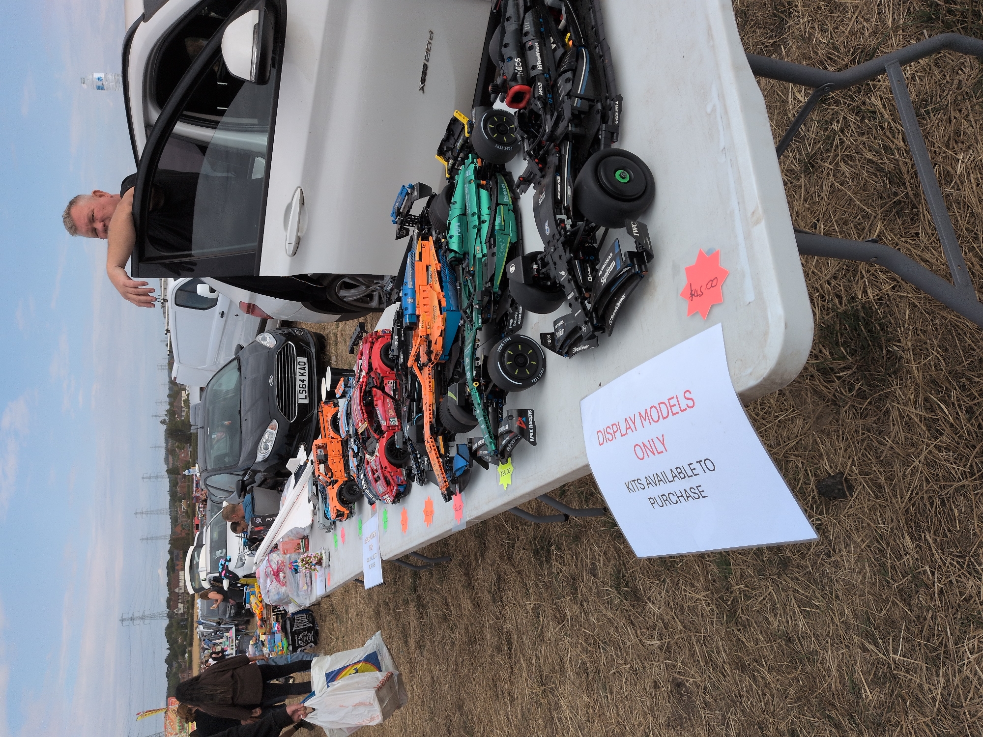 An outdoor market scene, likely a car boot sale or fete, is depicted. The foreground is dominated by a white folding table laden with an assortment of detailed model cars, appearing to be constructed from Lego. These models range in color, including black, green, orange, and red, and are arranged in rows. Some models have visible sponsor decals. A white sign, propped against the table's edge, reads "DISPLAY MODELS ONLY KITS AVAILABLE TO PURCHASE".
Behind the table, a white vehicle with its passenger door open is parked. A man, with short gray hair and wearing a dark t-shirt, leans casually against the car door. The background reveals a mix of parked cars, tents, and people browsing. In the distance, power pylons and a hazy sky suggest a semi-rural or suburban setting, consistent with the general area of South Staffordshire, United Kingdom, known for its numerous villages and open spaces where such events are commonly held. The ground is covered in dry grass and straw, indicating a potentially dry or autumnal day.