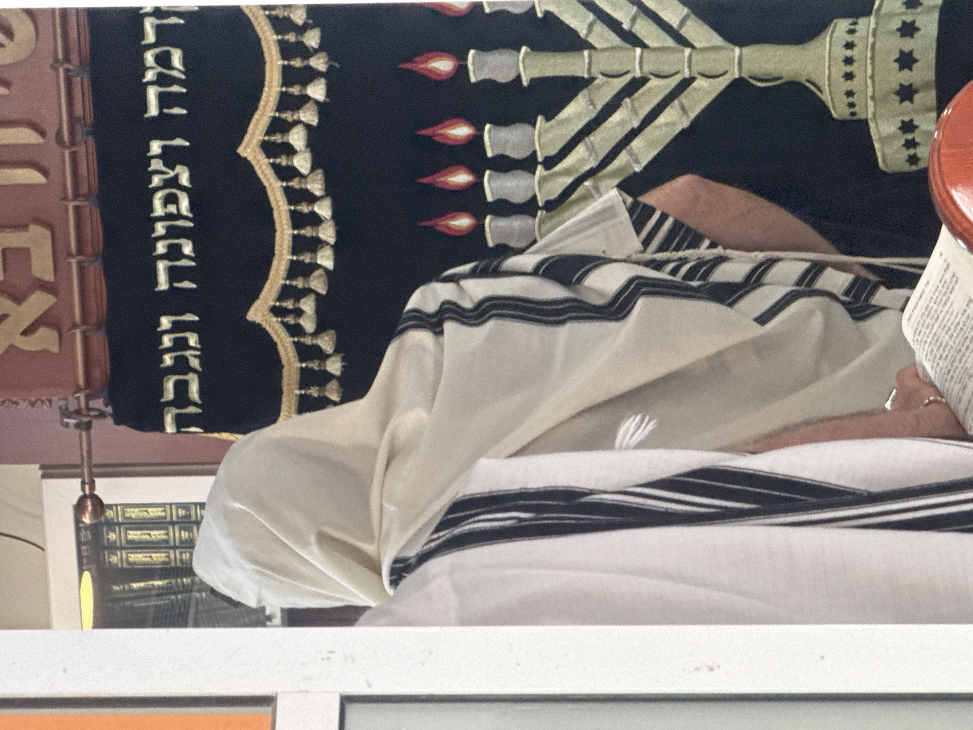 A photograph taken in Givatayim, Israel, depicts a person draped in a white tallit with black stripes. The person is likely engaged in religious practice. Behind the person, a black cloth features a menorah design. Gold Hebrew lettering is visible on the left side of the cloth. The backdrop includes what appears to be a bookshelf with books and a copper colored hanging object, possibly part of the synagogue's interior. The scene is indoors, probably within a synagogue.
