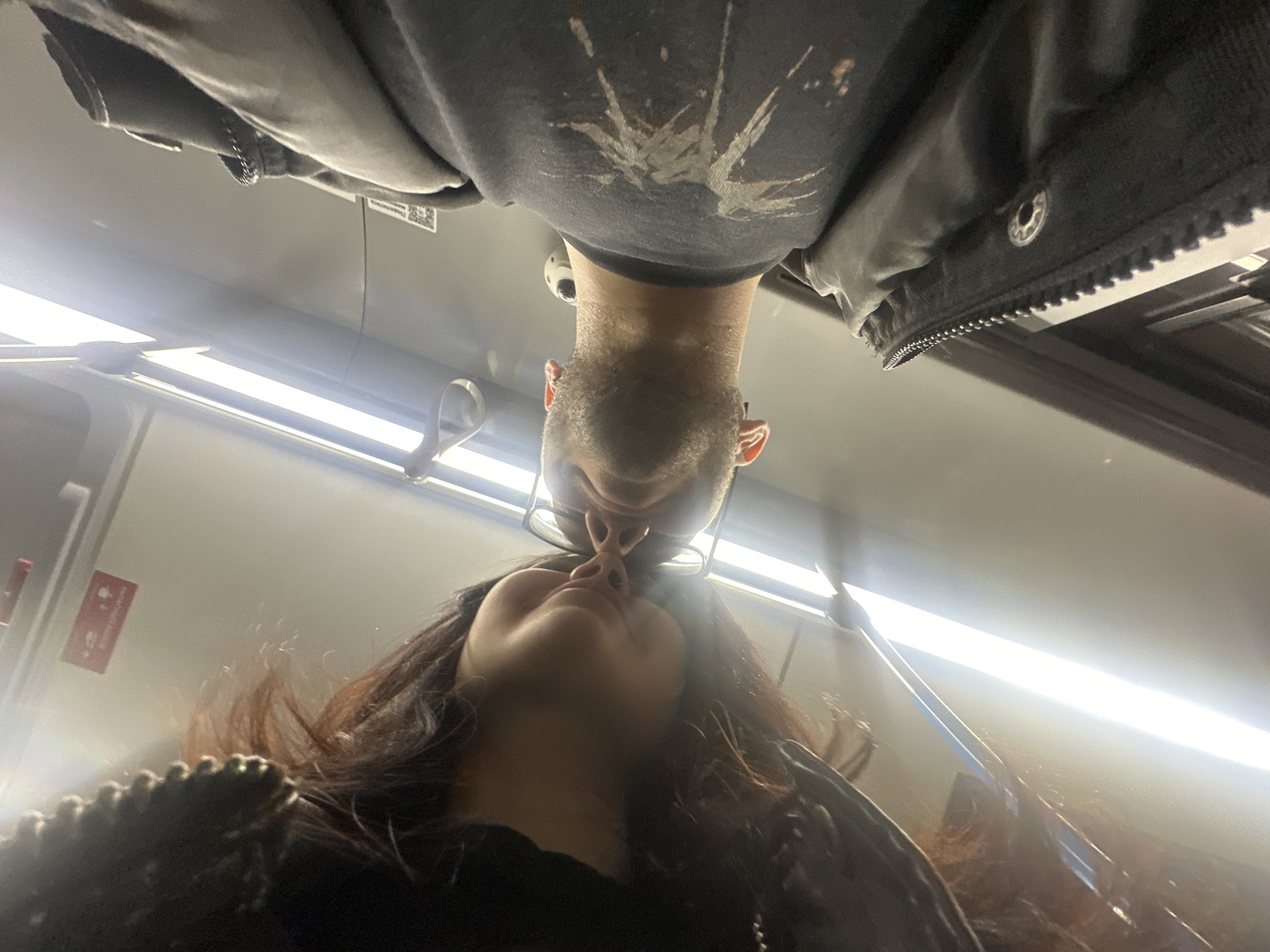 An inverted image shows two individuals, a male and a female, in an intimate pose, captured from a low-angle perspective inside what appears to be a public transport vehicle, likely a train or subway car, in Khimki, Russia. The male is positioned upside down, with his head slightly lower than his body, wearing glasses, a dark t-shirt with a light-colored design, and a dark jacket. He has short facial hair. The female is positioned below him, also upside down, looking upwards with long dark hair, wearing dark clothing. Their noses are touching, suggesting a tender, playful, or romantic moment.
The setting is characterized by a bright, white overhead interior with prominent fluorescent strip lighting running along the ceiling. Metal handles and bars are visible, typical of a public transport environment. A small security camera is discreetly visible in the background, above the male's head.
Notable details include a white tag with a QR code attached to an overhead compartment or luggage rack. Russian text is partially visible on this tag, reading "Пользователя" (Pol'zovatelya), meaning "User." To the left, a red sign with white text is visible on the wall; partially decipherable text includes "НЕ" (NE), meaning "DO NOT," which is often part of common signage like "Do not lean" in Russian transport. The artificial lighting suggests the time of day is not explicitly conveyed, and the indoor setting means weather is not a factor. The overall emotion conveyed is one of affection and closeness between the two subjects.