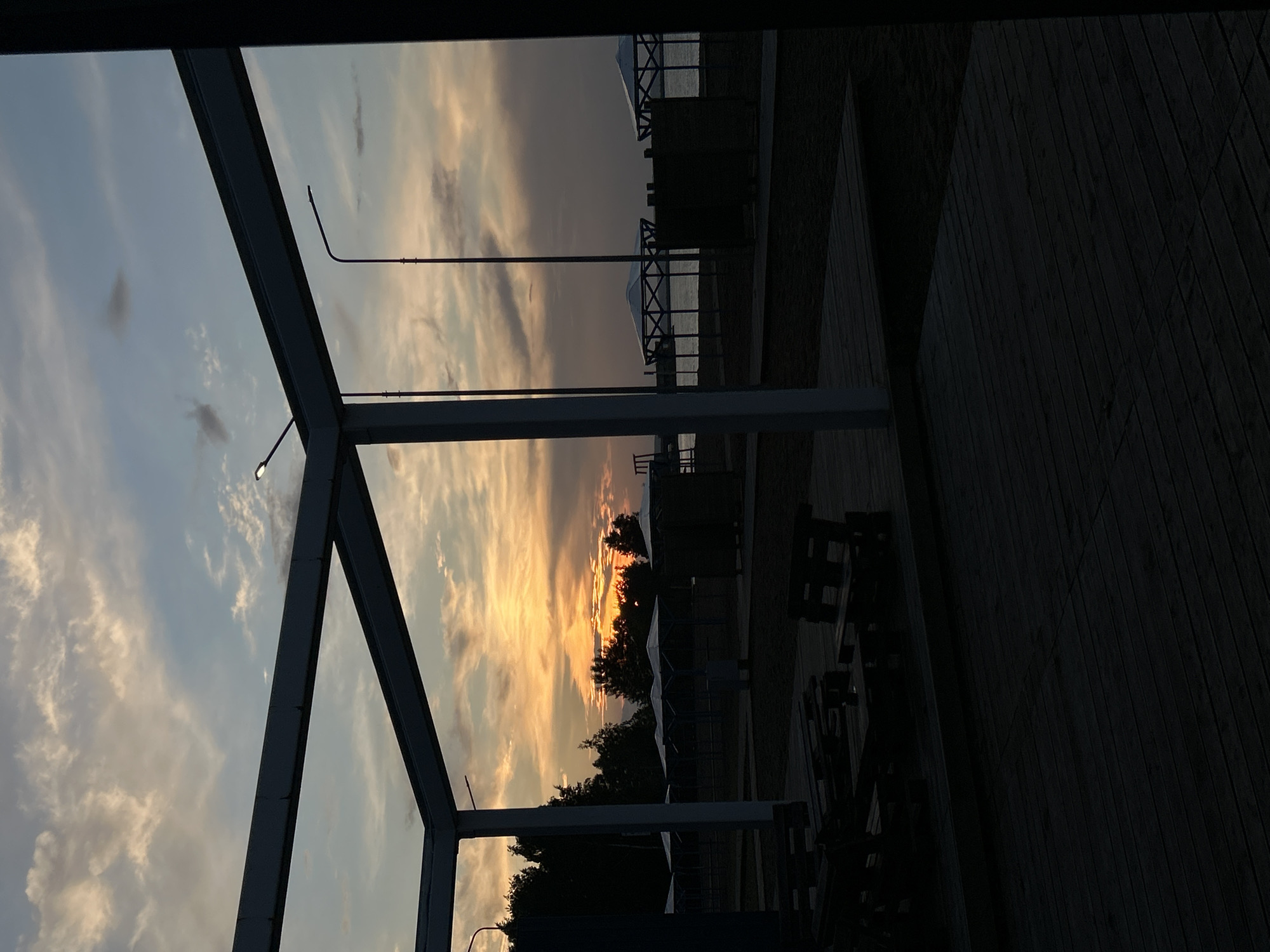 This image captures a serene sunset over the Kama River in Chaikovsky, Russia. The foreground is dominated by a dark wooden deck, with partially visible wooden benches and tables. A modern, white-framed structure with dark beams extends into the scene, framing the view. Beyond this structure lies a beach area with several white-capped gazebos. The water of the Kama River stretches across the middle ground, reflecting the soft hues of the twilight sky. The sky itself is a canvas of pale blue and scattered clouds, tinged with orange and yellow from the setting sun. The silhouette of trees lines the horizon behind the gazebos, adding depth to the landscape. Two streetlights are visible, one near the top left and another more prominently in the middle right, their lights unlit. The overall atmosphere is peaceful and tranquil, suggesting the end of a day.