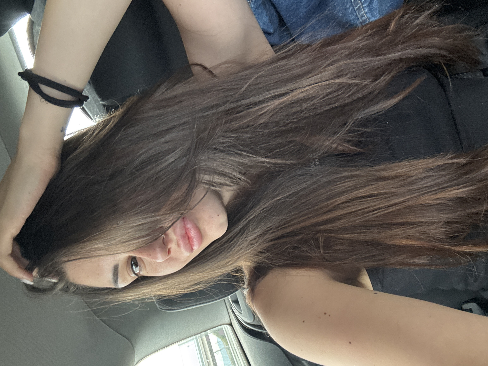 The image depicts a young woman inside a car, seen from a slightly high angle and rotated, giving a dynamic feel. Her long, voluminous dark brown hair, with subtle lighter brown highlights, dominates a large portion of the frame, flowing over her shoulders and chest.
She is looking towards the camera, with her left eye clearly visible, dark and engaging. Her nose and lips, slightly parted in a soft pout, are also in view. Her left arm is bent, with her hand near her head, and a black scrunchie is worn around her left wrist. A small, faint mark or tattoo is visible on her left forearm. She appears to be wearing a black top or garment, with a piece of blue denim fabric visible in the upper left background, possibly part of her attire or a jacket.
The setting is clearly the interior of a car, with elements like the headliner and blurred car window reflections visible. The lighting suggests it is daytime, with bright ambient light illuminating the scene. The overall impression is a casual portrait taken within a vehicle. There is no clearly discernible text within the image. The location context provided is Aldomirovtsi, Bulgaria.