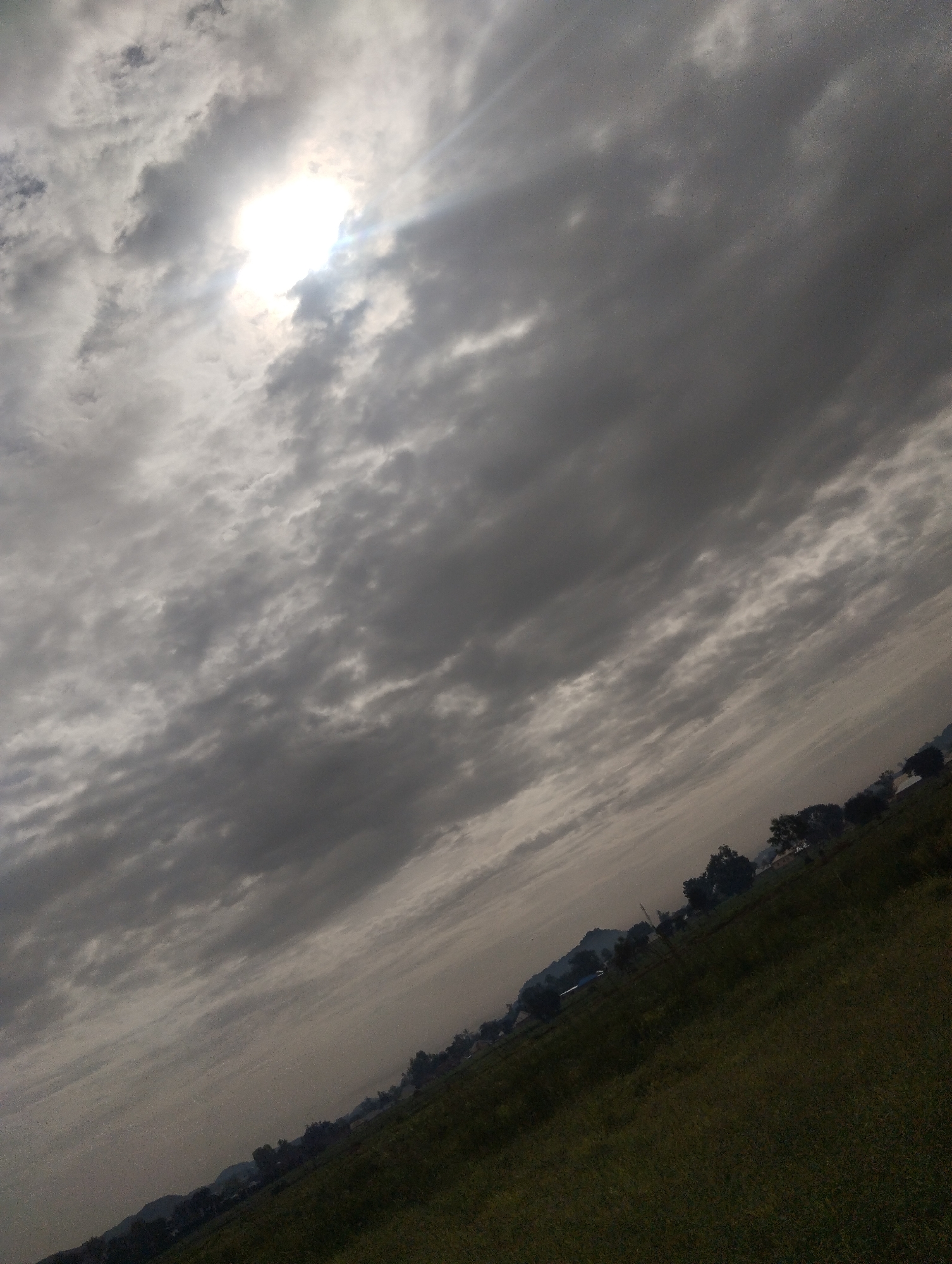 This media file depicts a landscape scene under a cloudy sky. The primary subjects are the sky filled with dramatic clouds, with the sun peeking through a bright opening, casting rays of light. Below the sky is a sloping landscape with green vegetation, appearing to be a field or hillside. Scattered trees and some structures, possibly houses or farm buildings, are visible along the horizon, suggesting a rural or semi-rural setting. The overall mood is somewhat somber due to the heavy clouds, but the sunlight breaking through adds a touch of hope or drama. The scene is likely taking place in the afternoon or late afternoon, given the angle of the sun and the cloud cover. There are no people or specific objects that are clearly identifiable as central to an activity. The location cues, such as the vegetation and type of structures, are consistent with a setting like Bukuru, Nigeria. No visible text is present in the image.
