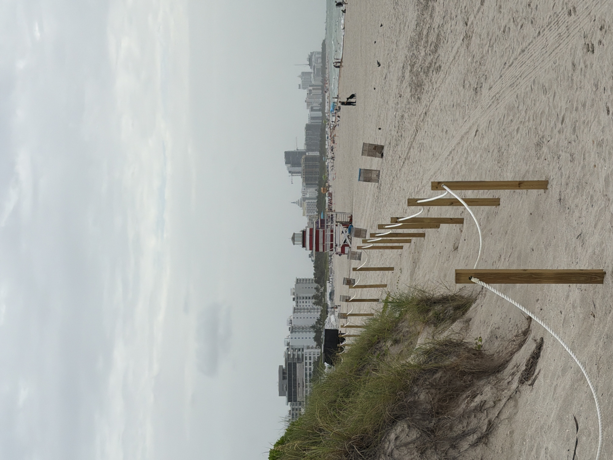 An overcast day at South Beach in Miami Beach, Florida. A rope barrier, supported by wooden posts, guides people along the sand towards the ocean. In the background, a line of high-rise buildings forms the Miami skyline. People are scattered across the beach, some sunbathing, others walking or playing. A lifeguard stand, painted in red and white stripes, stands prominently on the sand. The sand itself is a pale beige, with tire tracks visible from maintenance vehicles. The overall atmosphere is one of a typical beach day, albeit under cloudy skies.