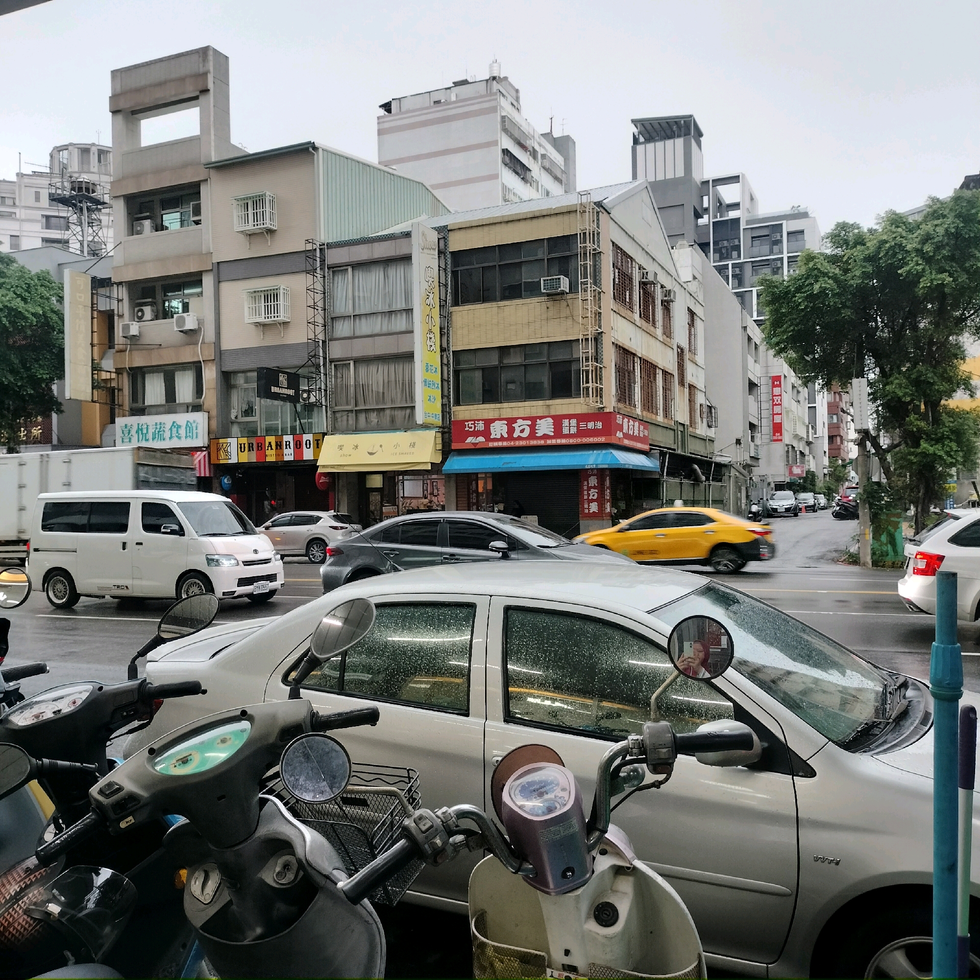 The image captures a street scene in Taichung, Taiwan, on a cloudy, possibly rainy day. The foreground is dominated by two parked scooters. Behind them, a silver sedan is parked, its windows streaked with rain. Further back, a white van drives down the wet street, followed by a yellow car. Buildings line the street, displaying various architectural styles and signage in Chinese characters, indicating commercial establishments such as restaurants and shops. The sky is overcast, and the overall impression is one of a typical urban environment in Taiwan.