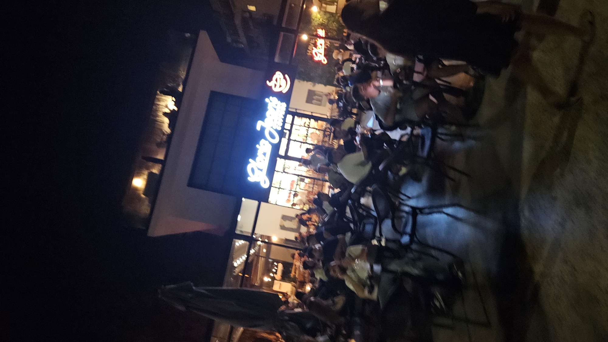 This image depicts a nighttime scene at a Gloria Jean's Coffees establishment in Fethiye, Turkey. A considerable number of patrons are seated at tables and chairs outside the cafe. The building facade is illuminated with neon signs, with "Gloria Jean's Coffees" prominently displayed in cursive script. Red neon lights also spell out "HAVE Gloria Jean's". The ground appears to be paved. Several individuals are visible, some seated, others walking, and engaging in conversations. The atmosphere suggests a bustling evening at a popular cafe.