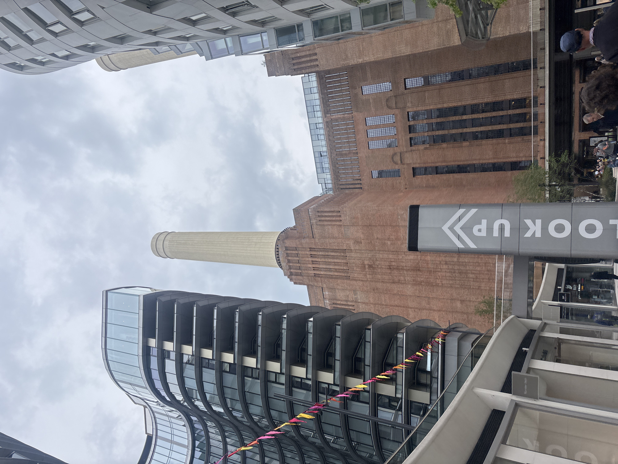This image shows the Battersea Power Station in London, United Kingdom, viewed from a low angle. The sky is overcast.
The power station, a large brick building, is the dominant structure. To the left, there is a modern building with curved balconies. A string of colorful pennants hangs from the modern building. To the right, the edge of another building is visible. A sign with the words "LOOK UP" is in the foreground, partially obscuring the view of the power station. People are walking near the power station entrance.