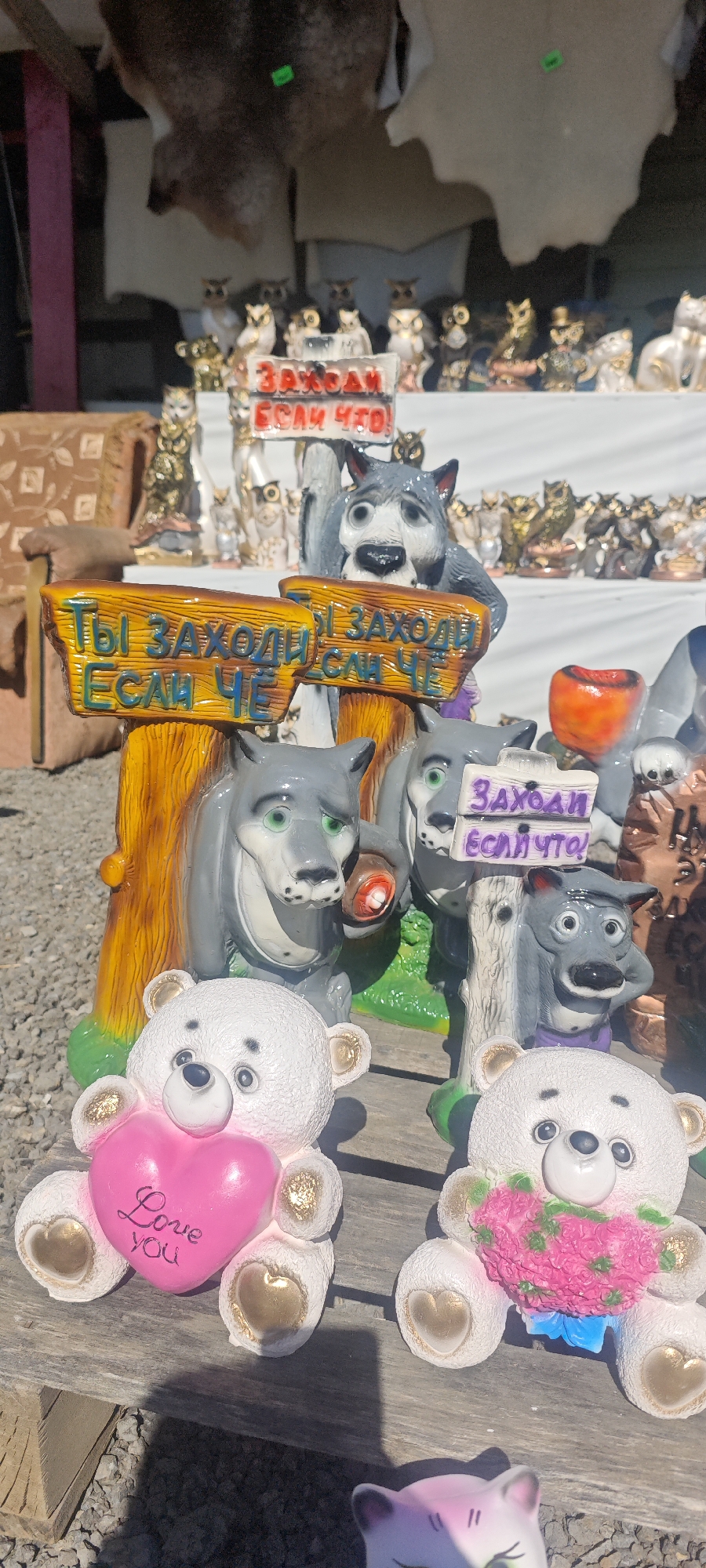 The image shows a display of ceramic figurines for sale. In the foreground are two white teddy bears, one holding a pink heart with the words "Love You" and the other holding pink flowers with a blue bow. Behind the bears are ceramic wolf figurines, some holding signs. The signs are in Russian and roughly translate to "Come in, just in case". In the background, more ceramic figures, including owls and cats, are visible. The scene is set outdoors, possibly at a market. The location is speculated to be in the city of Olen'e, Russia.