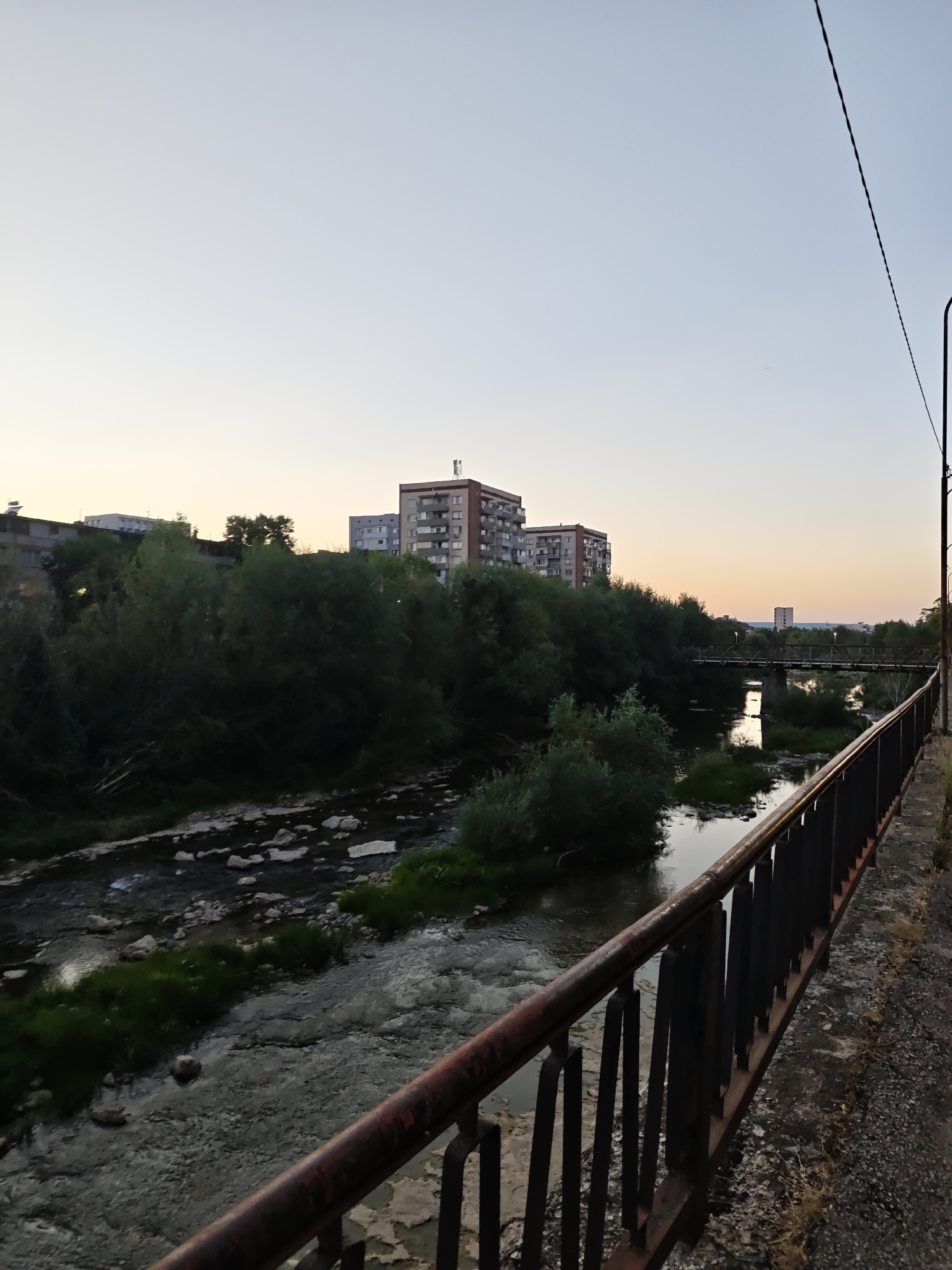 The image depicts a river flowing through an urban environment, likely in Lovech, Bulgaria. The foreground features a weathered railing with rusty accents, bordering a concrete path. Beyond the railing, the river is visible, with rocky beds and patches of green grass along its banks. Trees and dense foliage line the river, creating a natural barrier between the water and the city structures in the background. Several apartment buildings are visible, some with visible antennas. A pedestrian bridge spans the river in the middle ground. The sky is a pale blue with a gradient of yellow and orange near the horizon, suggesting either dawn or dusk. A utility pole with a wire stretches across the upper right portion of the frame. The scene is devoid of people or any discernible activity.