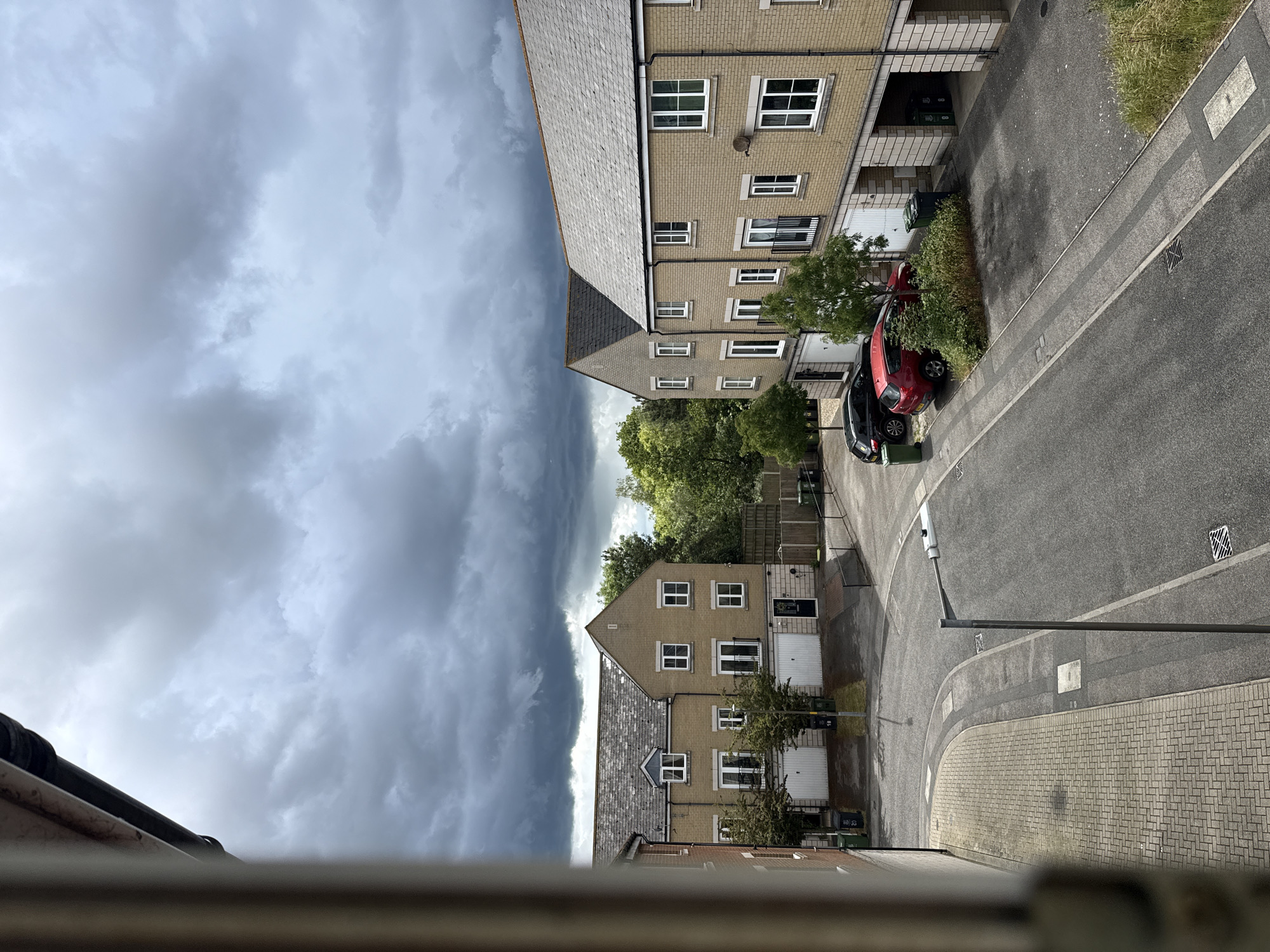 The image captures a residential street scene under a dramatic, heavily clouded sky. The photograph appears to be taken from an elevated position, looking down and across at the street and opposing houses.
The sky dominates the upper portion of the frame, filled with dark, dense grey clouds, suggesting an impending storm or heavy rain. The clouds have a textured, rolling appearance, with lighter grey patches indicating some breaks or varied density.
Below the sky, on the right side of the image (if oriented vertically), are several two-story houses. They feature light-colored brick or rendered facades, white window frames, and grey slate-tiled roofs. Some houses have integrated garages with white doors. Trees and green bushes are visible along the frontages of these houses and between properties. A red car is parked on what appears to be a driveway or verge, partially obscured by greenery, with a dark-colored car parked behind it.
The street itself is asphalt with paved sections, including a pedestrian pathway made of light grey block paving. A streetlamp stands in the middle distance. Green wheelie bins are visible near some of the houses and parked cars. The general architecture and residential layout are characteristic of a modern housing development in the United Kingdom, specifically Great Yarmouth as per the context.
The foreground shows a blurry railing or window ledge, indicating the viewpoint. No discernible human activity or explicit text is visible in the frame. The overall atmosphere is moody due to the overcast and stormy sky.